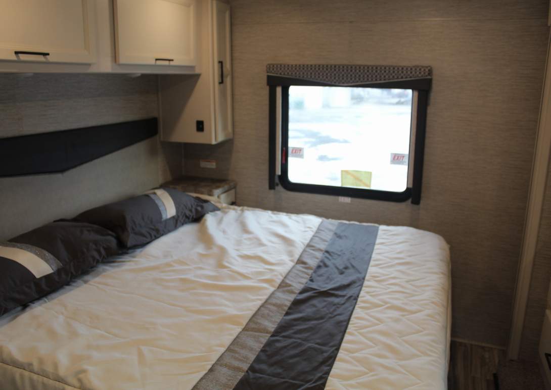 Inside an RV rental, a cozy bedroom featuring a neatly made bed, dark pillows, white overhead cabinets, and a window view.. Thor Motor Coach ace 2023