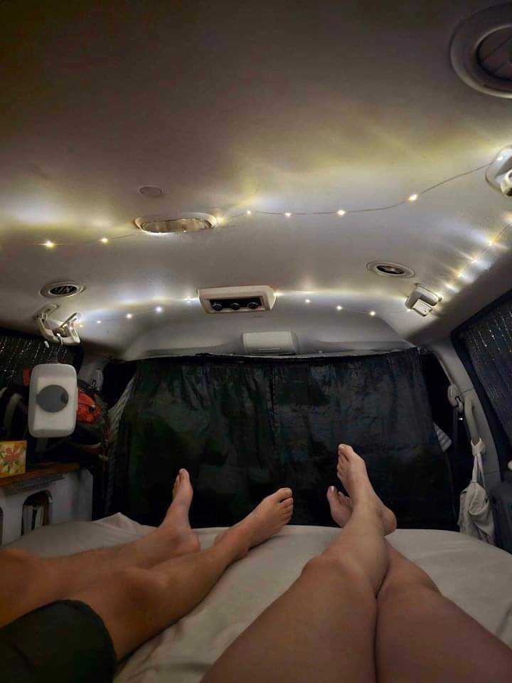 Relaxing in this cozy RV rental for van life. String lights illuminate two pairs of feet on the bed.. Dodge Grand Caravan 2009