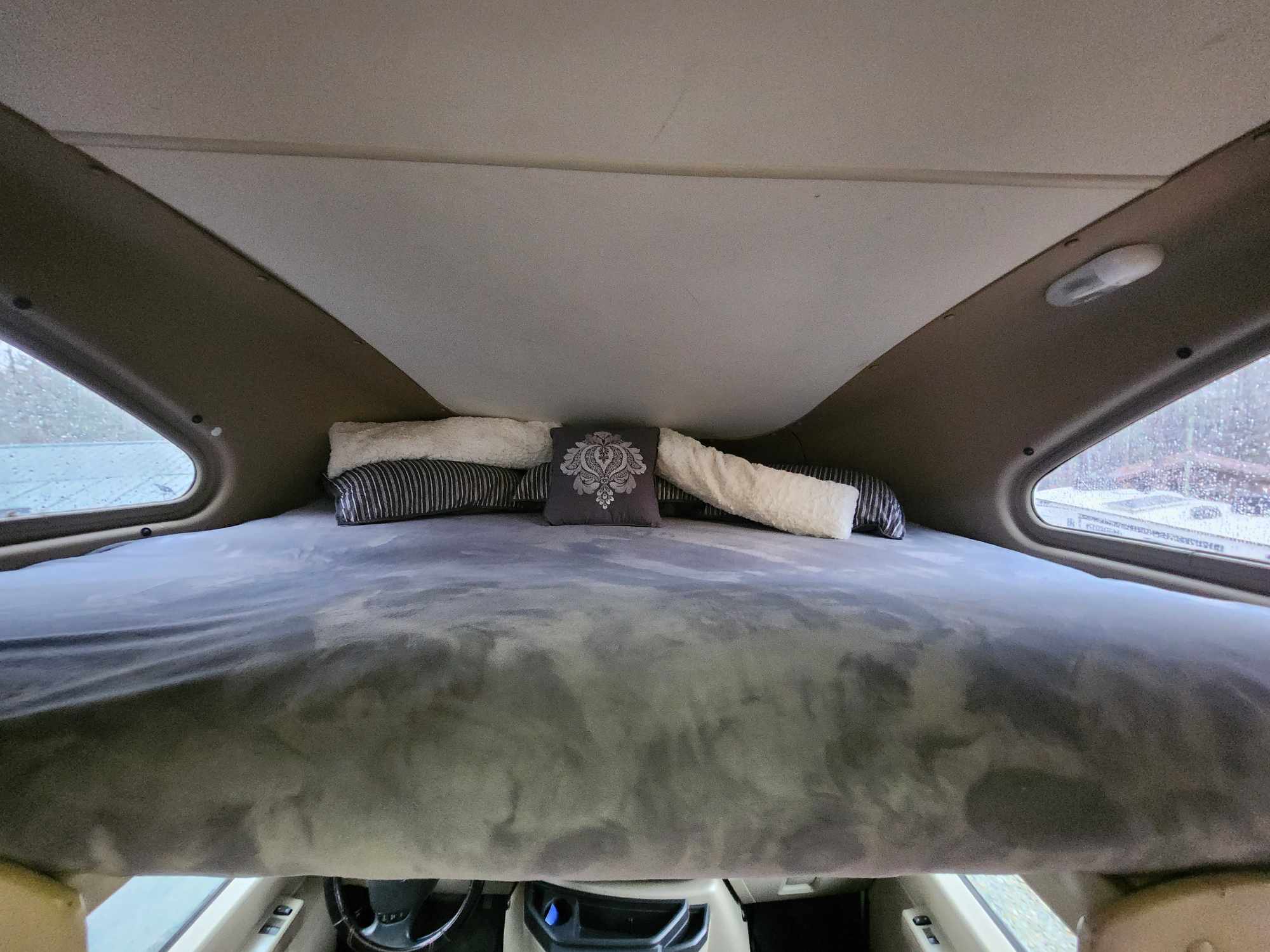 Inside an RV rental: cozy cab-over sleeping bunk with a plush grey bed and decorative pillows. View from above the driver's seat.. Thor 2012 Thor 19G 2012