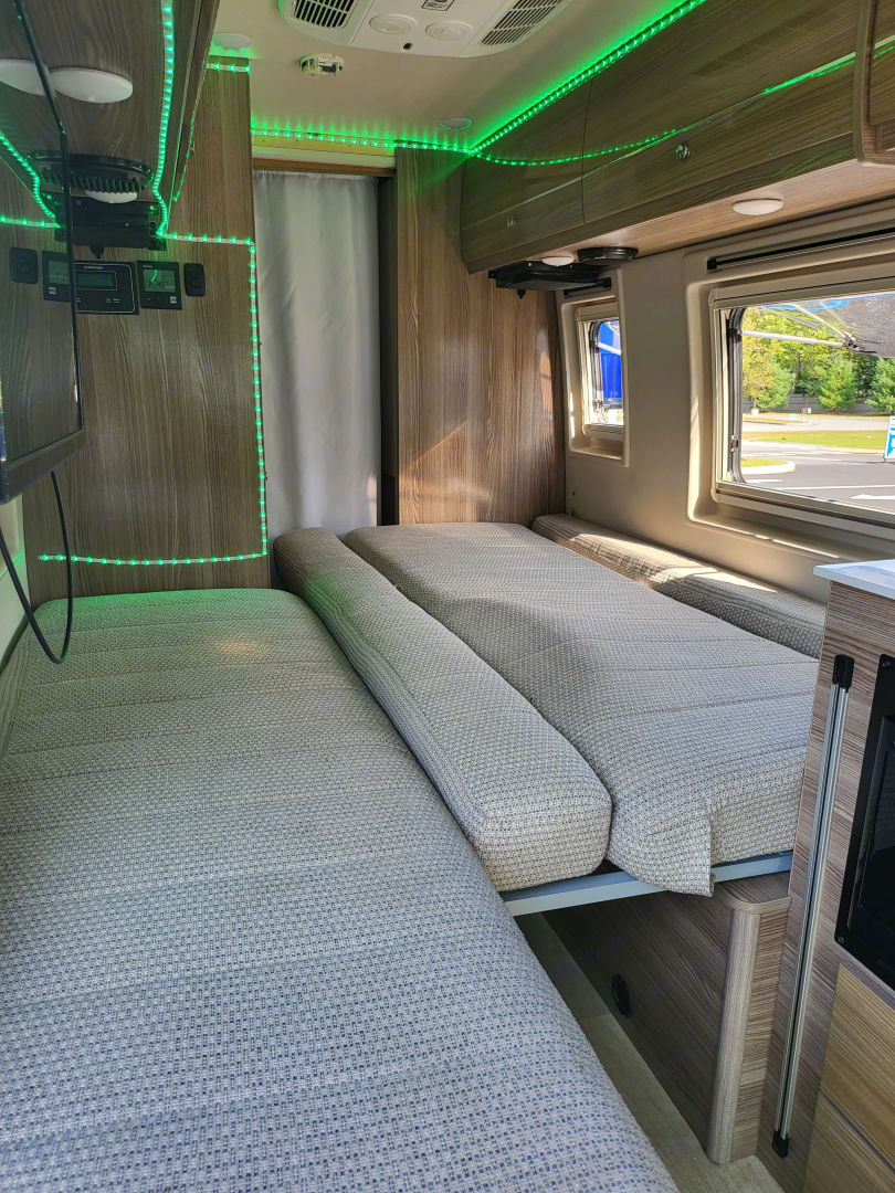 Modern RV rental interior: Wood-paneled walls, gray cushioned benches that form a bed, & green accent lighting.. Winnebago Boldt 2020