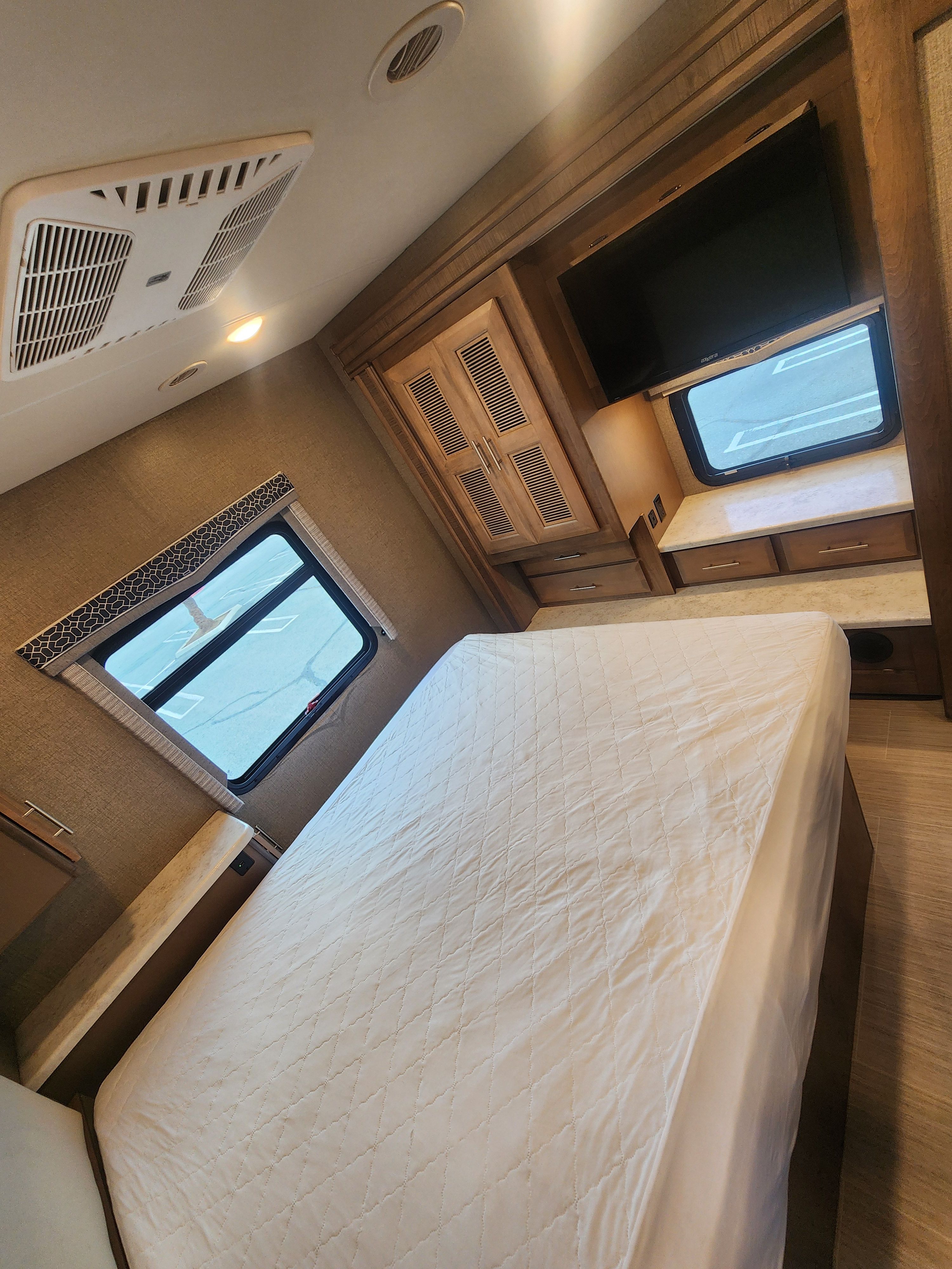 Interior of an RV rental bedroom featuring a large bed with white quilted cover, windows, mounted TV, and wood cabinetry.. Thor Motor Coach Magnitude 2021