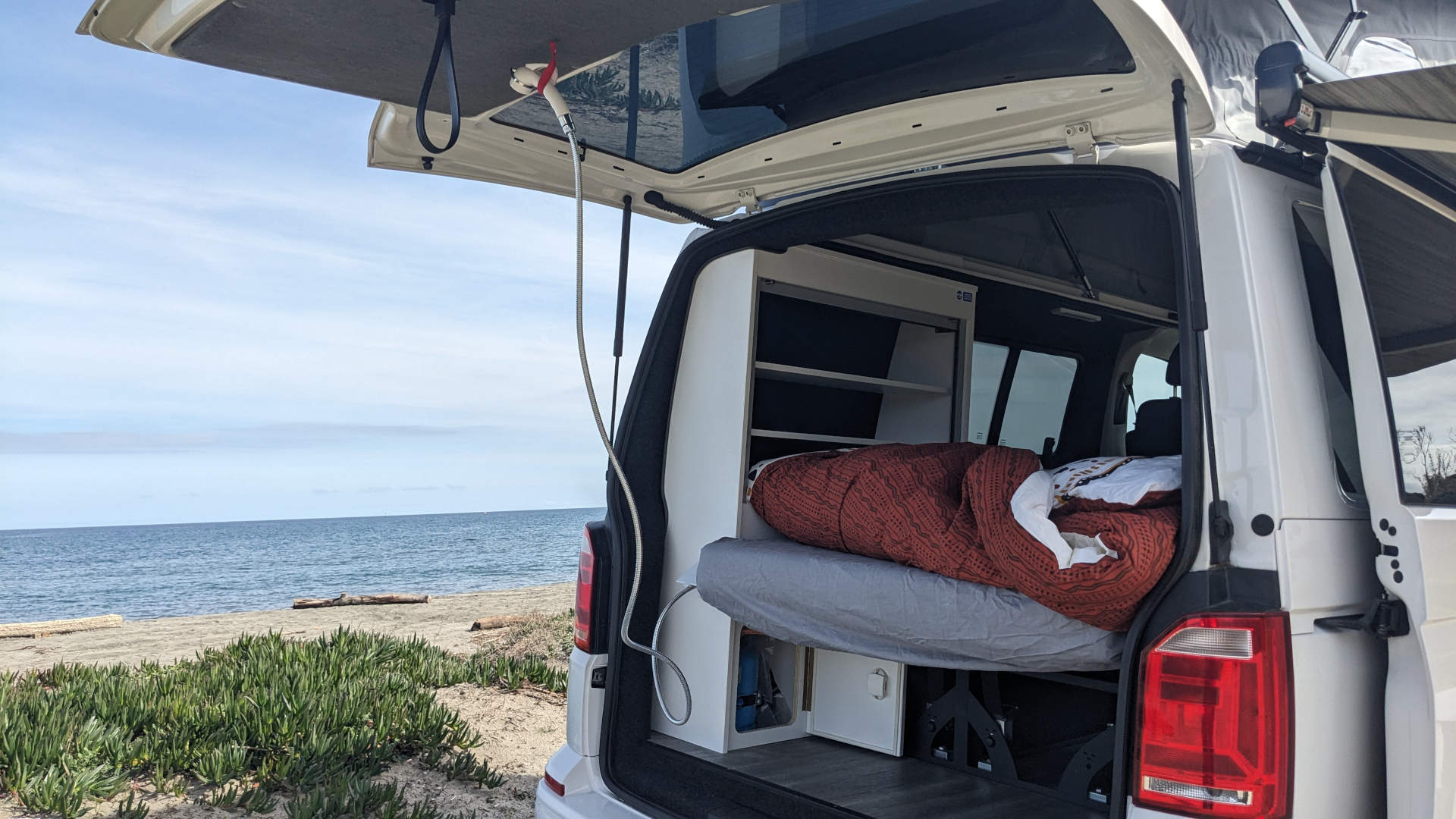 Cozy RV rental: Inside a camper van, a bed & outdoor shower face the ocean on a sandy beach.. Volkswagen T6 transporter 2019