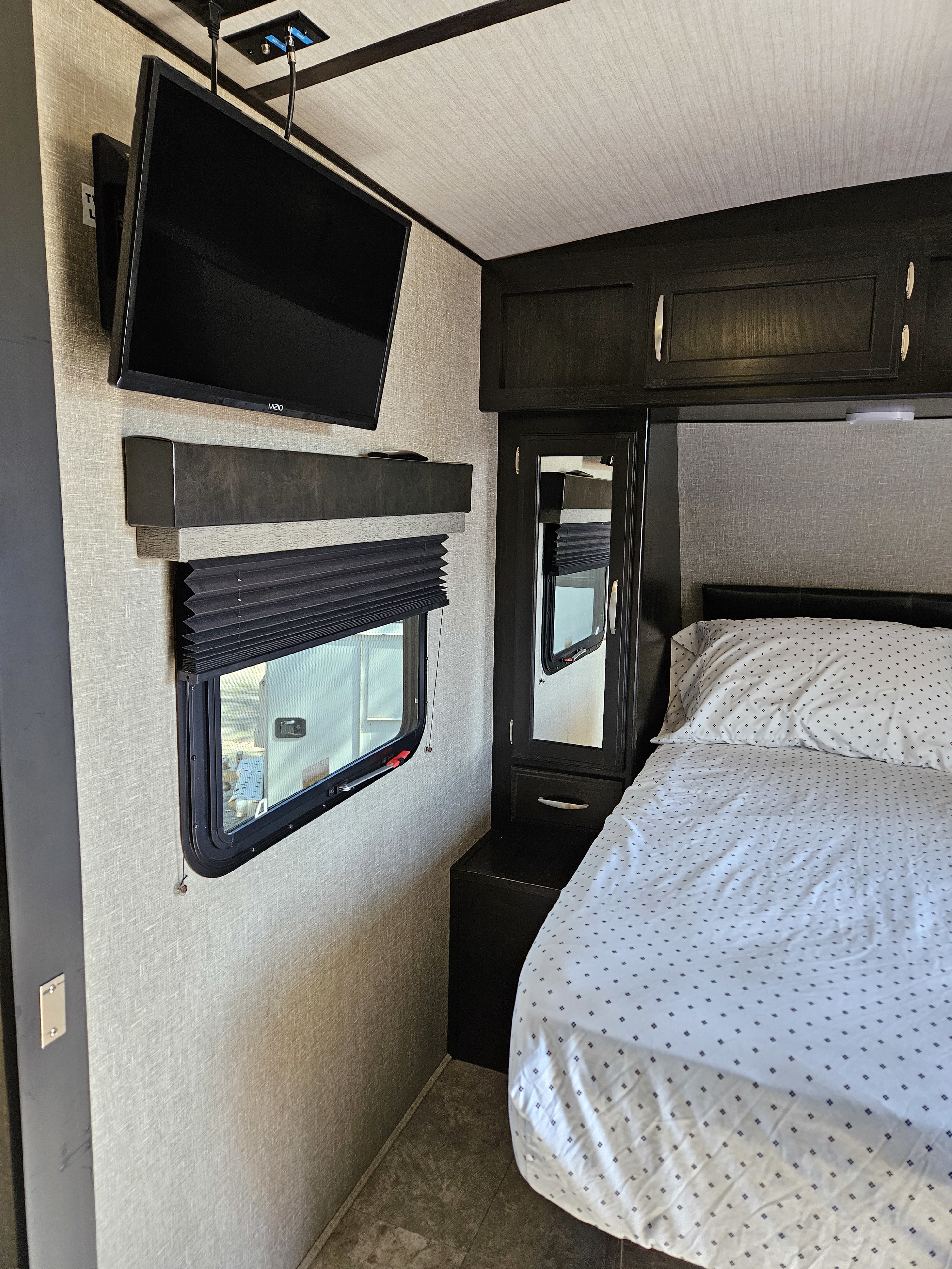 Cozy RV rental bedroom interior with wall-mounted TV, window, and comfortable bed. Dark wood cabinetry completes the space.. Grand Design Imagine 2019
