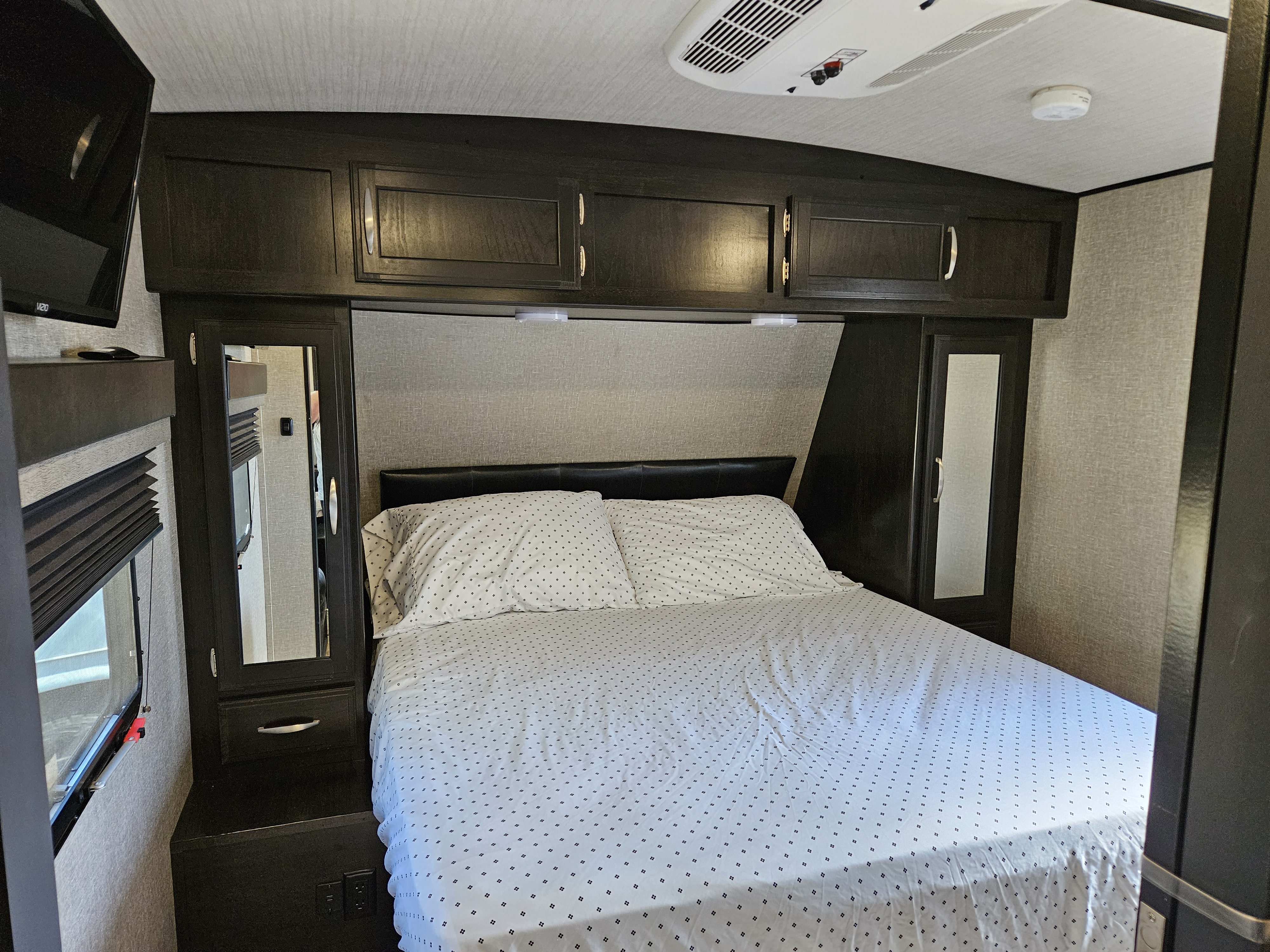 Comfortable bedroom in an RV rental, featuring a patterned bed, overhead cabinets, TV, and ceiling AC unit.. Grand Design Imagine 2019