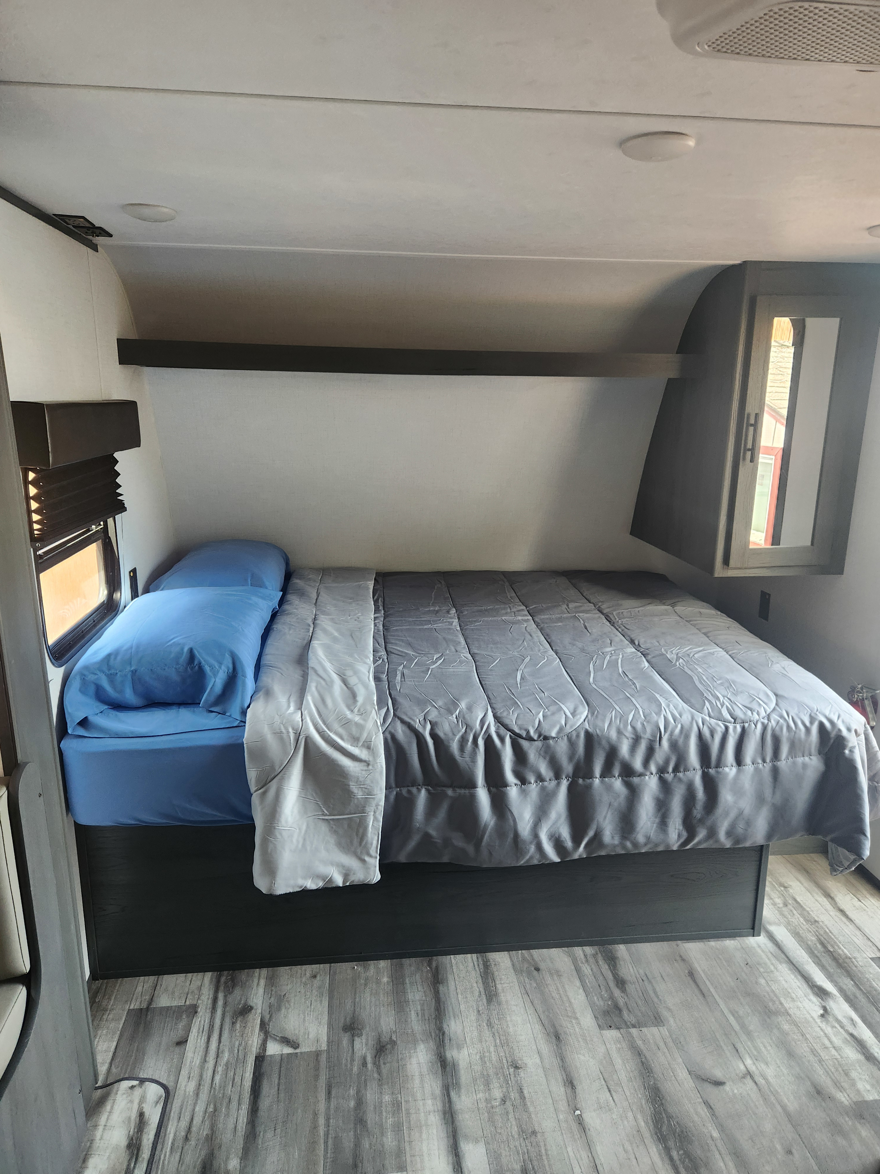 Cozy RV rental bedroom featuring a full bed with blue sheets & grey comforter, overhead storage, window, and wood-look flooring.. Forest River ASPEN TRAIL 2024