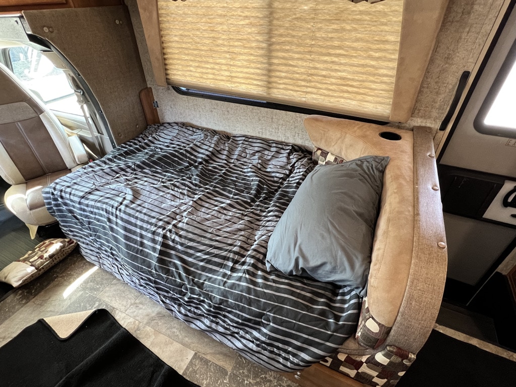 Experience comfort in our RV rental! This cozy interior features a bed with striped bedding next to the front cab.. Coachmen Leprechaun 2018