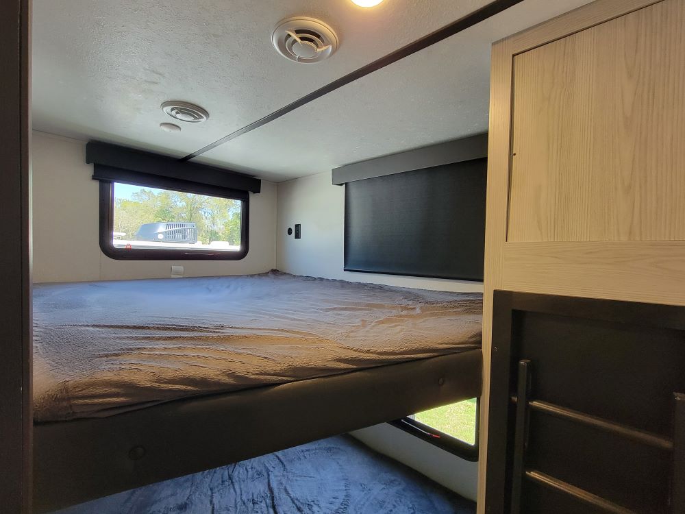 Inside an RV rental: a comfortable top bunk with a large window. Bottom bunk & storage visible.. Keystone RV Montana High Country 2022