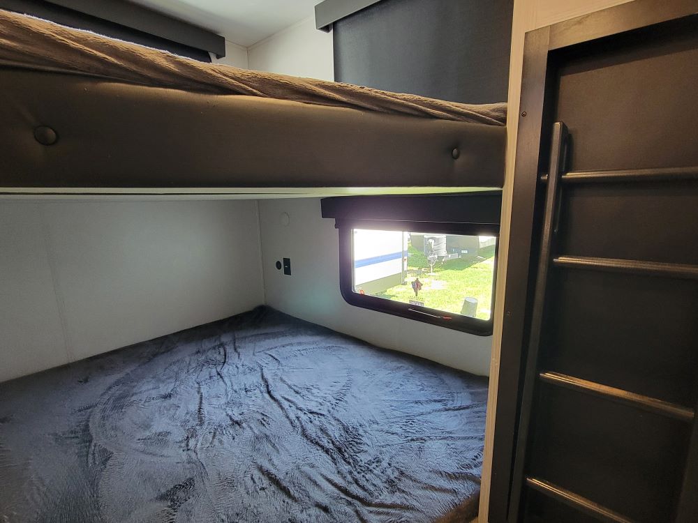 Inside an RV rental: upper & lower bunk beds with dark blankets. A window shows the outside, plus a ladder.. Keystone RV Montana High Country 2022