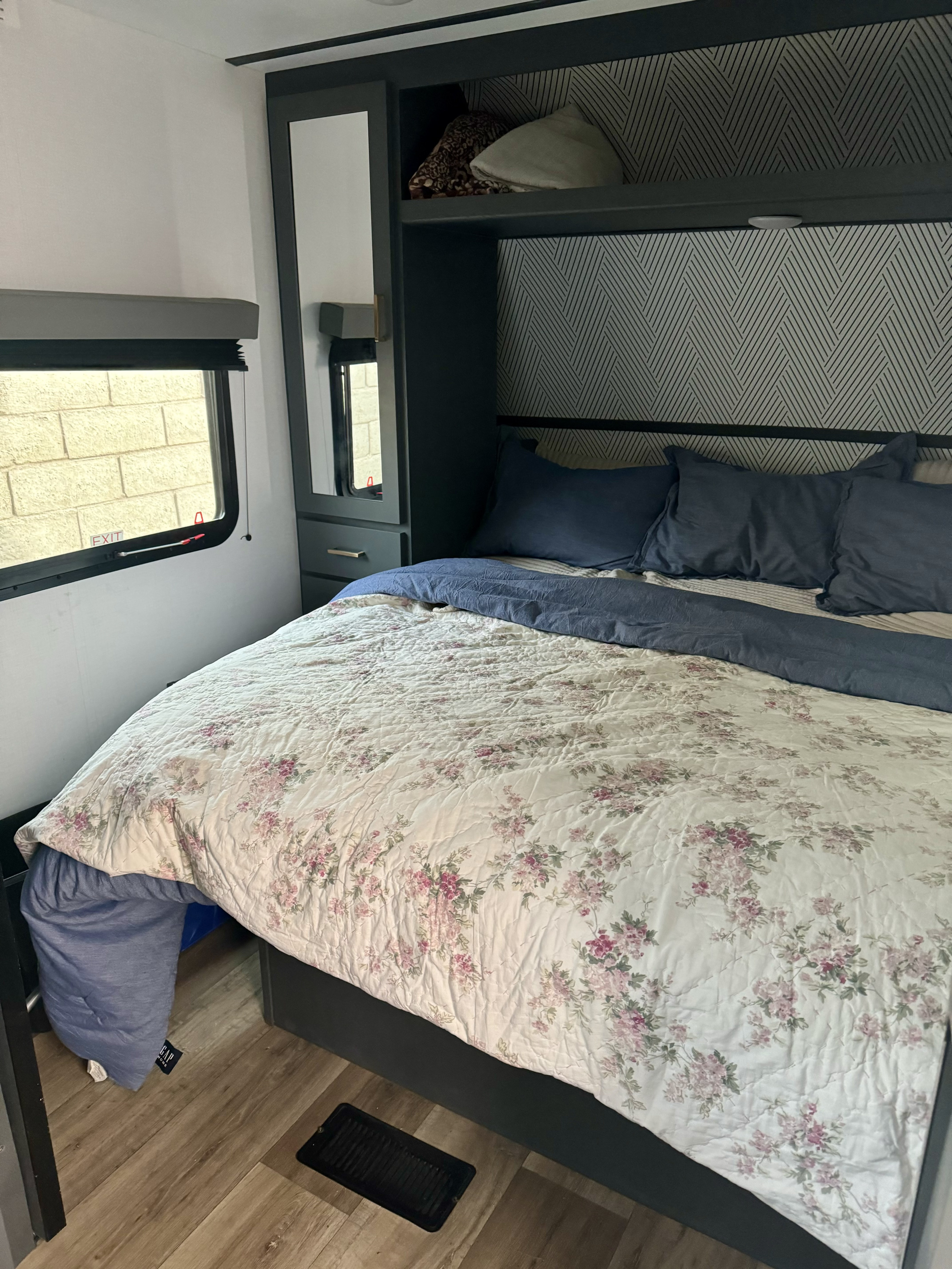 RV rental bedroom: floral bed, blue pillows, dark built-in storage with mirror, window, and patterned wall.. Keystone RV Bullet Crossfire 2022