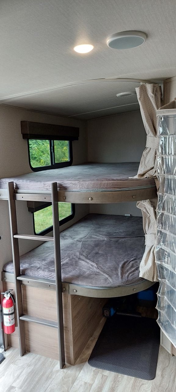 Inside an RV rental: two bunk beds with grey covers, a window, ladder, and fire extinguisher for a comfortable stay.. Grand Design Imagine 2021