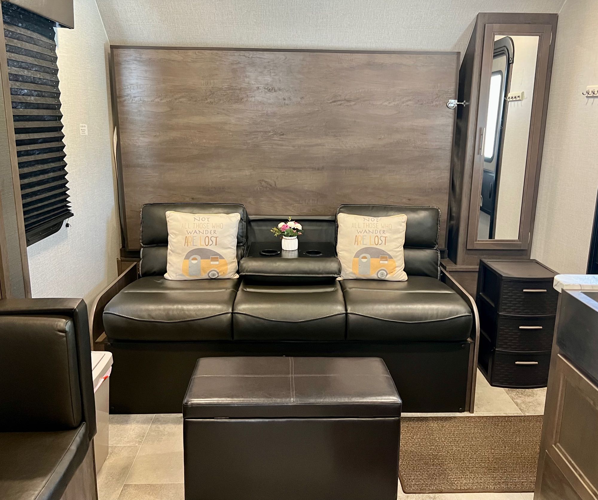 Compact RV rental living area with black leather sofa, camper pillows, and storage ottoman. Cozy, functional space for travel.. Dutchmen Coleman Light LX 2021