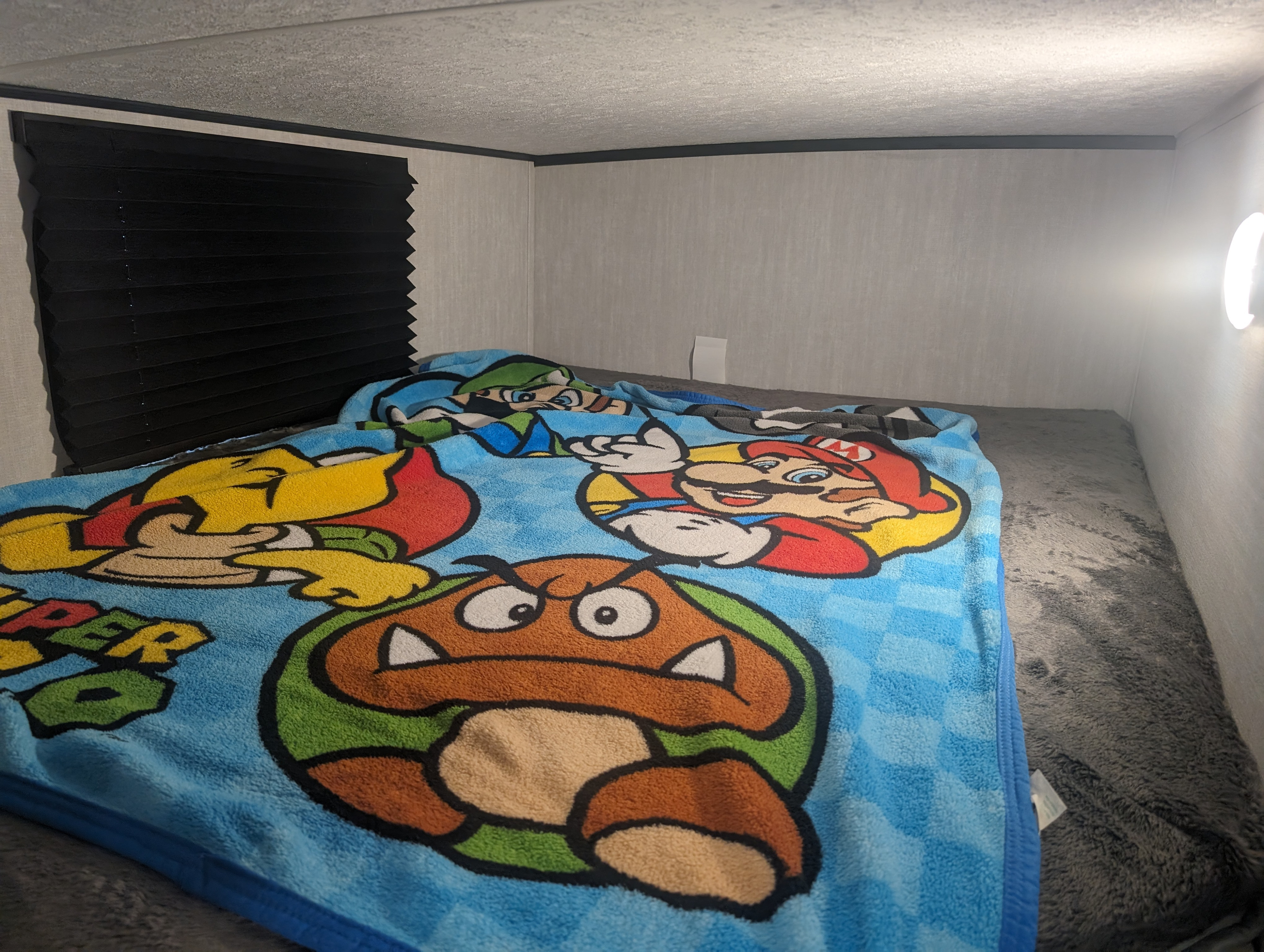 Cozy RV rental bedroom with a Super Mario blanket on the bed, black blinds, and a bright wall light.. Forest River Coachman Apex Nano 208BHS 2024