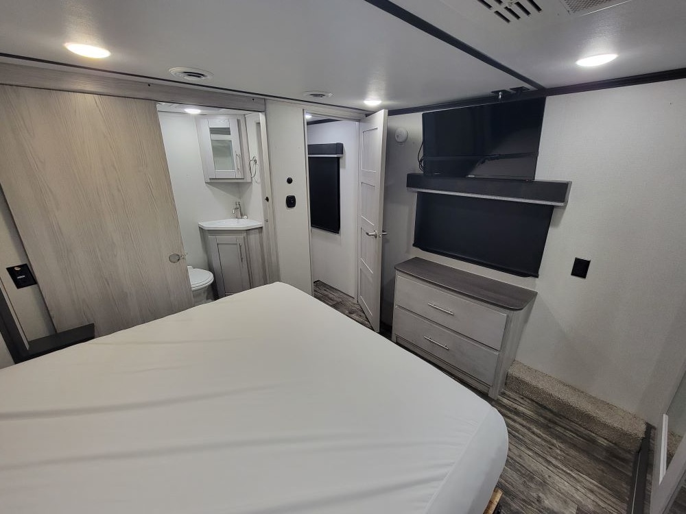 RV rental bedroom: features a comfy bed, mounted TV, dresser, and compact en-suite bathroom on the left. Ready for your trip.. Keystone RV Fuzion Toy Hauler 2021