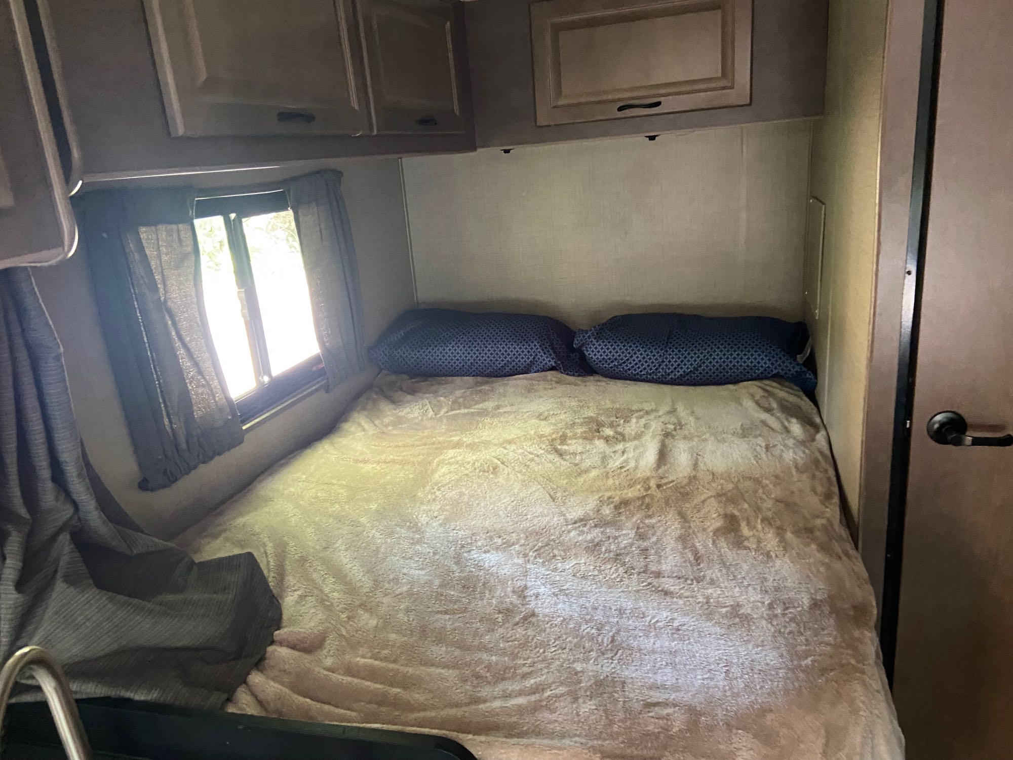 An RV rental bedroom, featuring a neatly made bed with two dark blue pillows, overhead cabinets, and a bright window.. Thor Majestic 2019