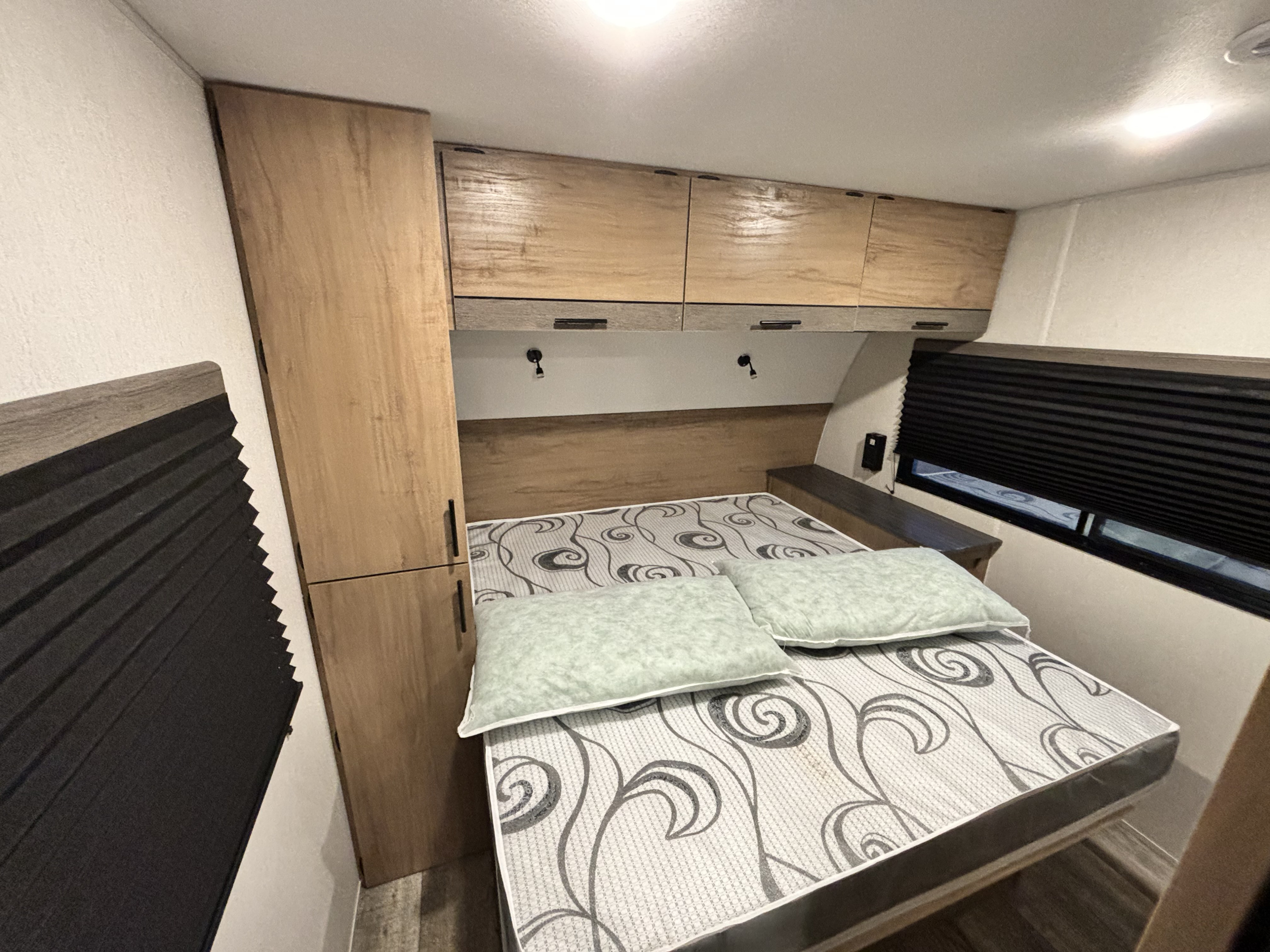 This rv rental bedroom features a comfortable bed, overhead cabinets, a tall wardrobe, and a window with blinds.. Forest River Campsite Reserve 2024