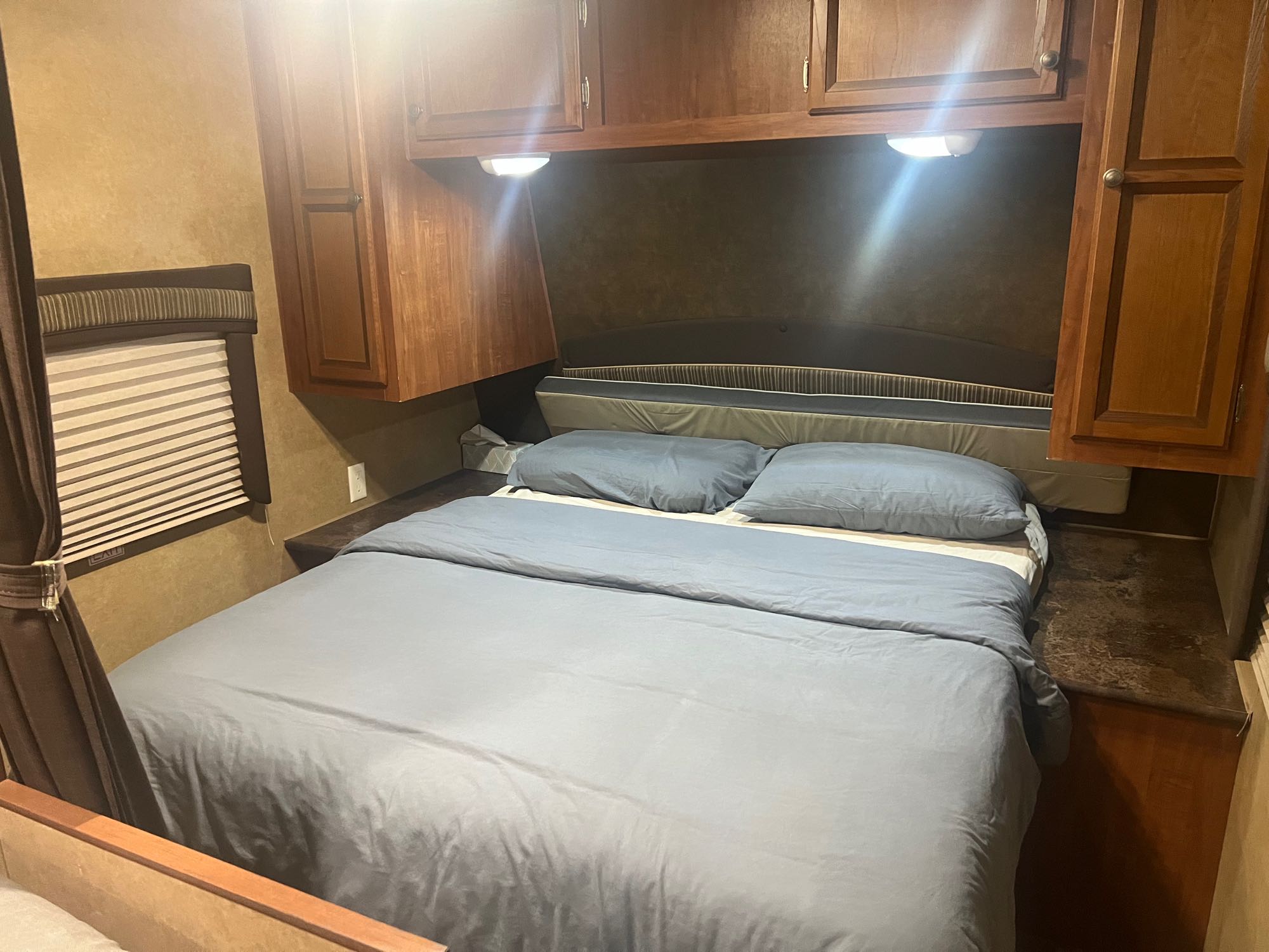 Cozy RV rental bedroom featuring a neatly made bed with blue bedding, overhead wooden cabinets, and bright lights.. Jayco Jay Flight 2013
