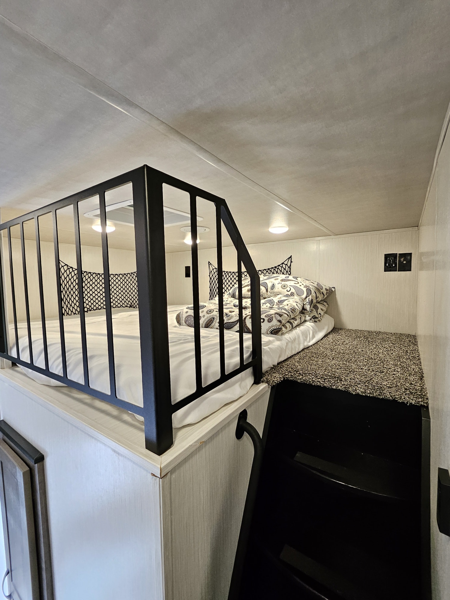 An RV rental's inviting loft bed, complete with a dark safety railing, patterned bedding, and carpeted floor leading to black stairs below.. Forest River Cedar Creek Fifth Wheel 2022