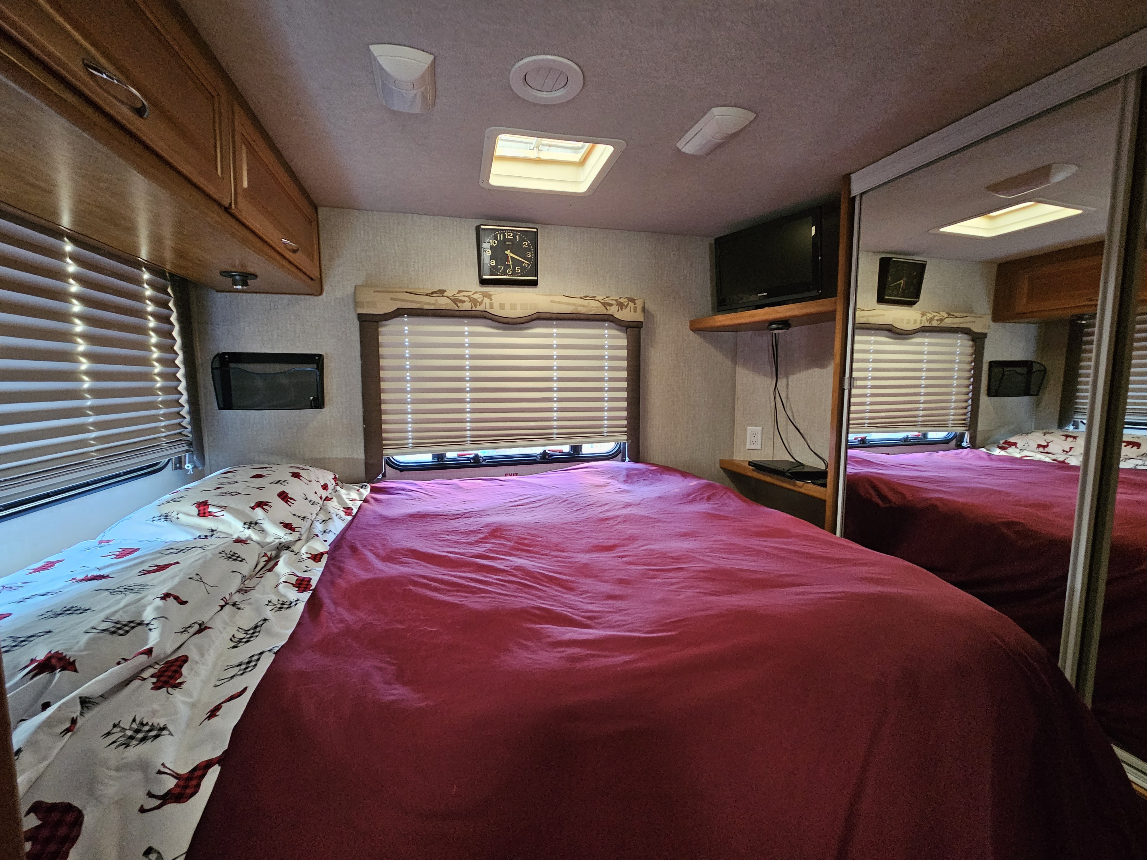 Inside a comfortable RV rental bedroom, featuring a vibrant red bed, window blinds, overhead cabinets, and a TV.. Tioga Tioga Motorhome 2008
