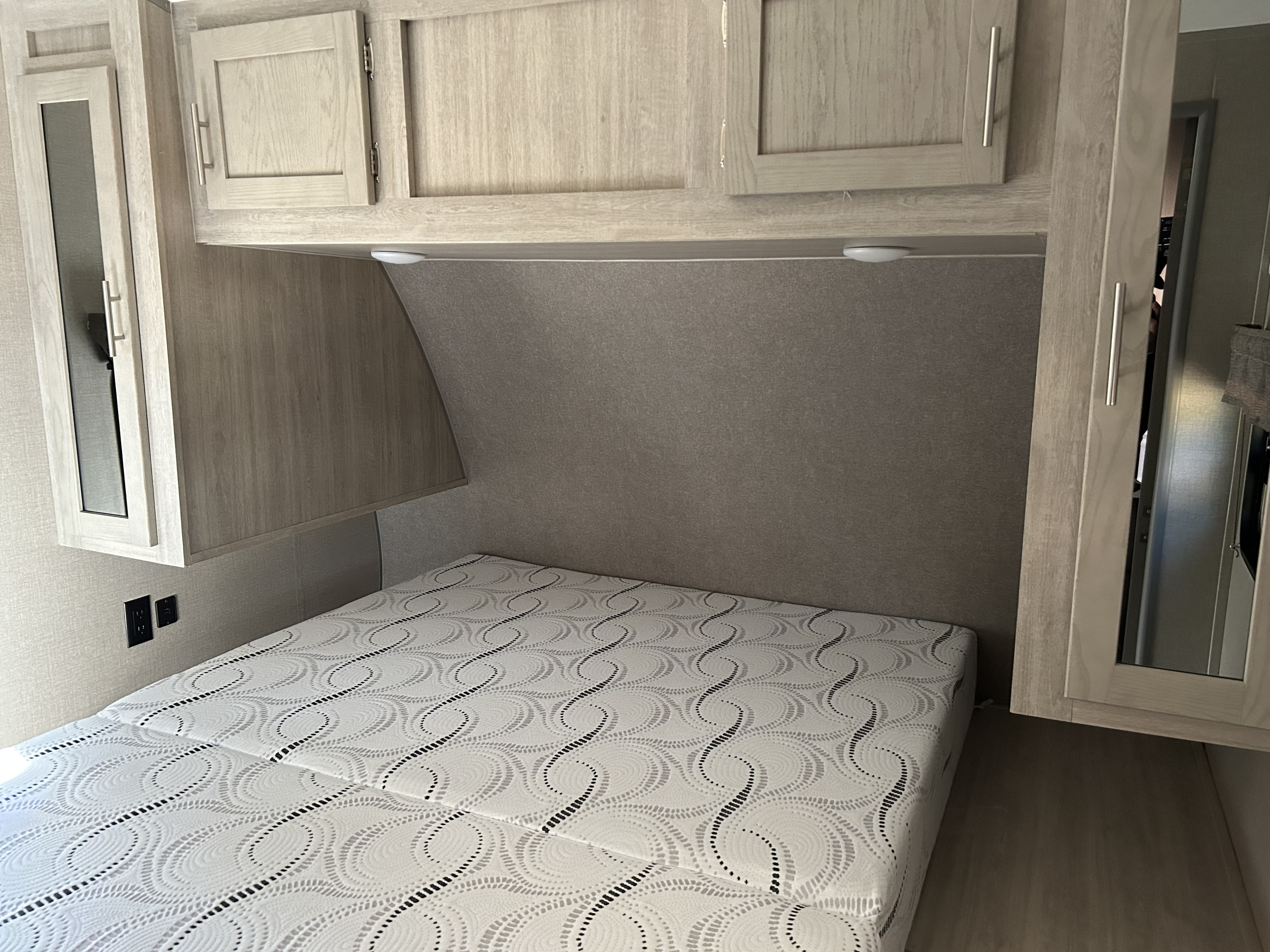 An RV rental's cozy bedroom, featuring a patterned mattress, light wood overhead cabinets, and essential wall outlets.. Coachmen Catalina Legacy 2022