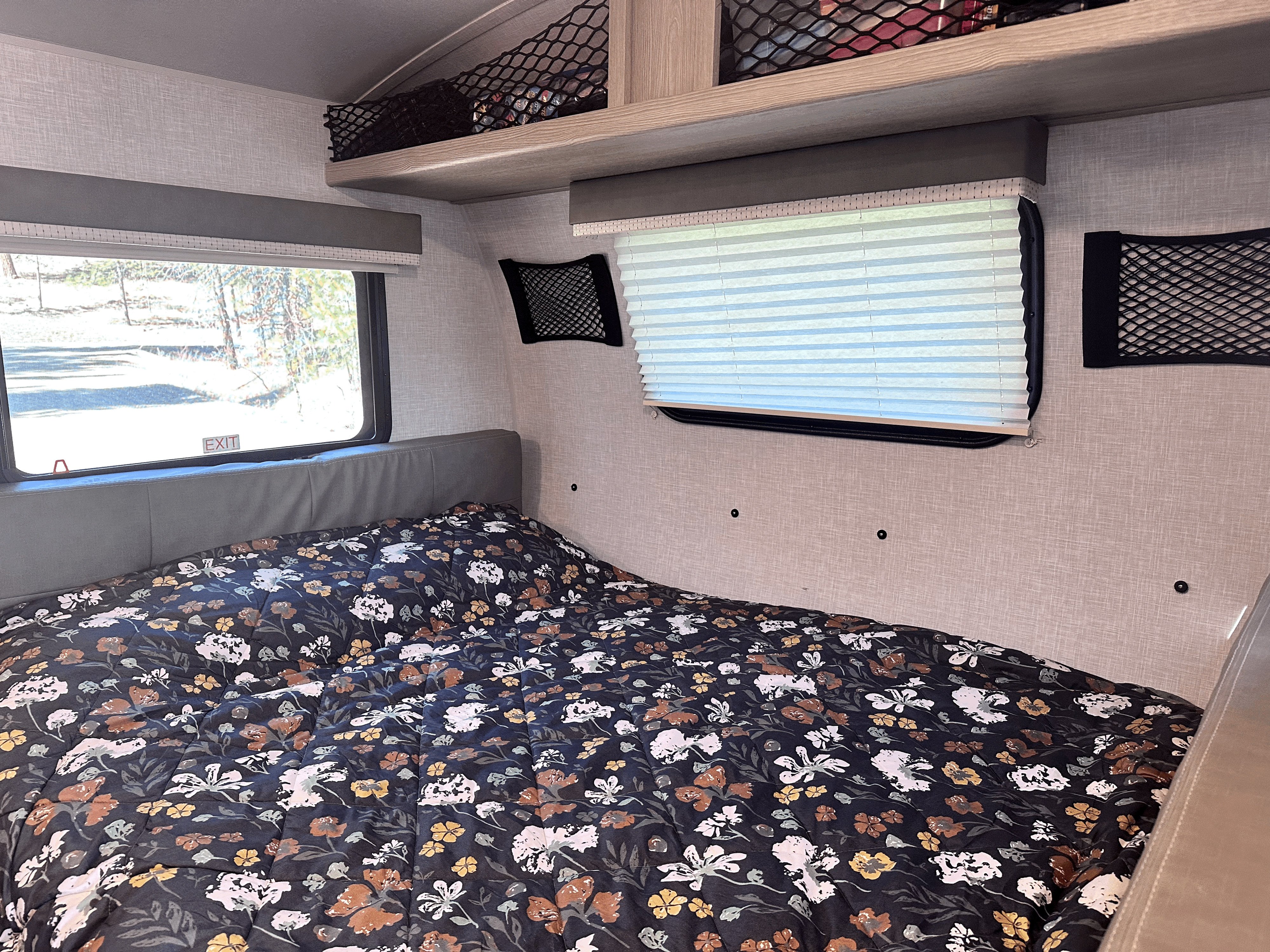 Cozy RV rental bedroom with a floral comforter, storage nets, and windows showing outdoor views. Perfect for your next trip!. Forest River Rockwood Geo Pro 2021