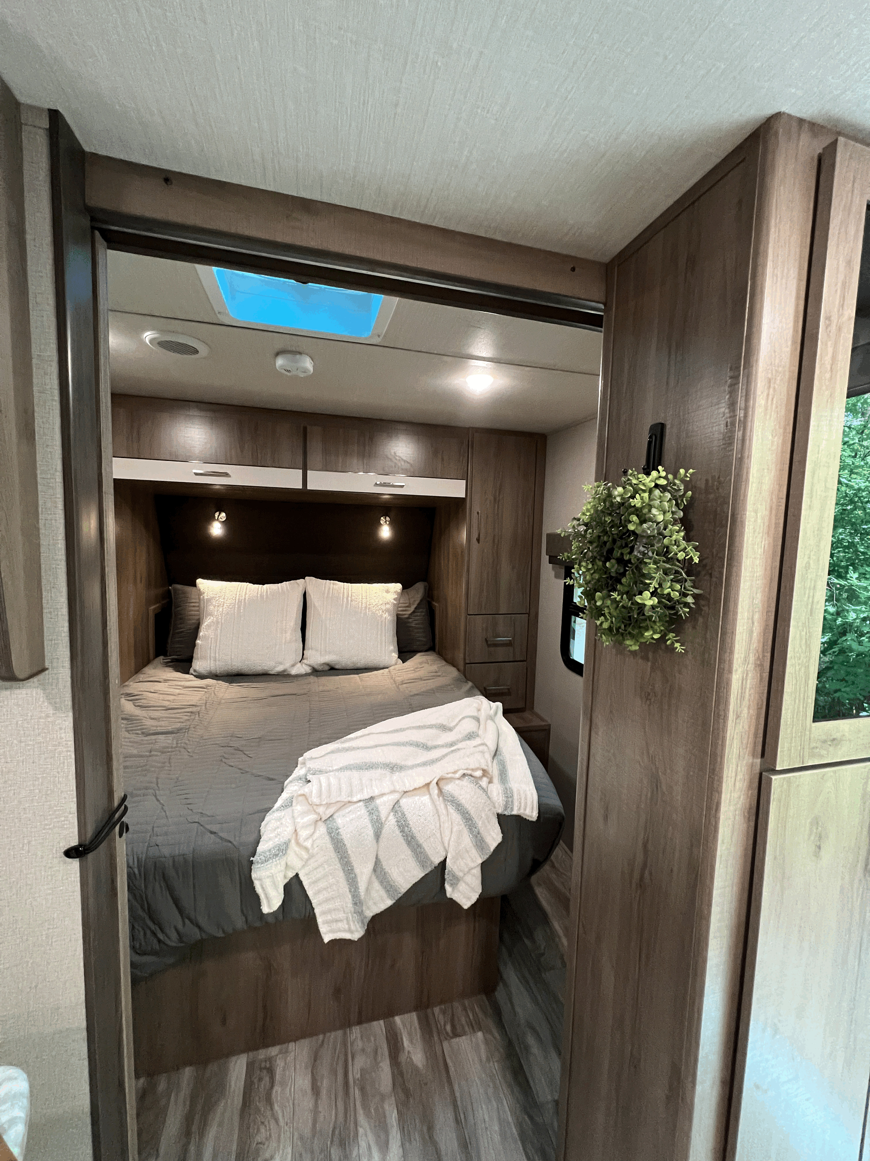 Modern RV bedroom in an RV rental, featuring a comfortable bed, wood-look cabinetry, and a bright skylight.. Grand Design Imagine XLS 2023