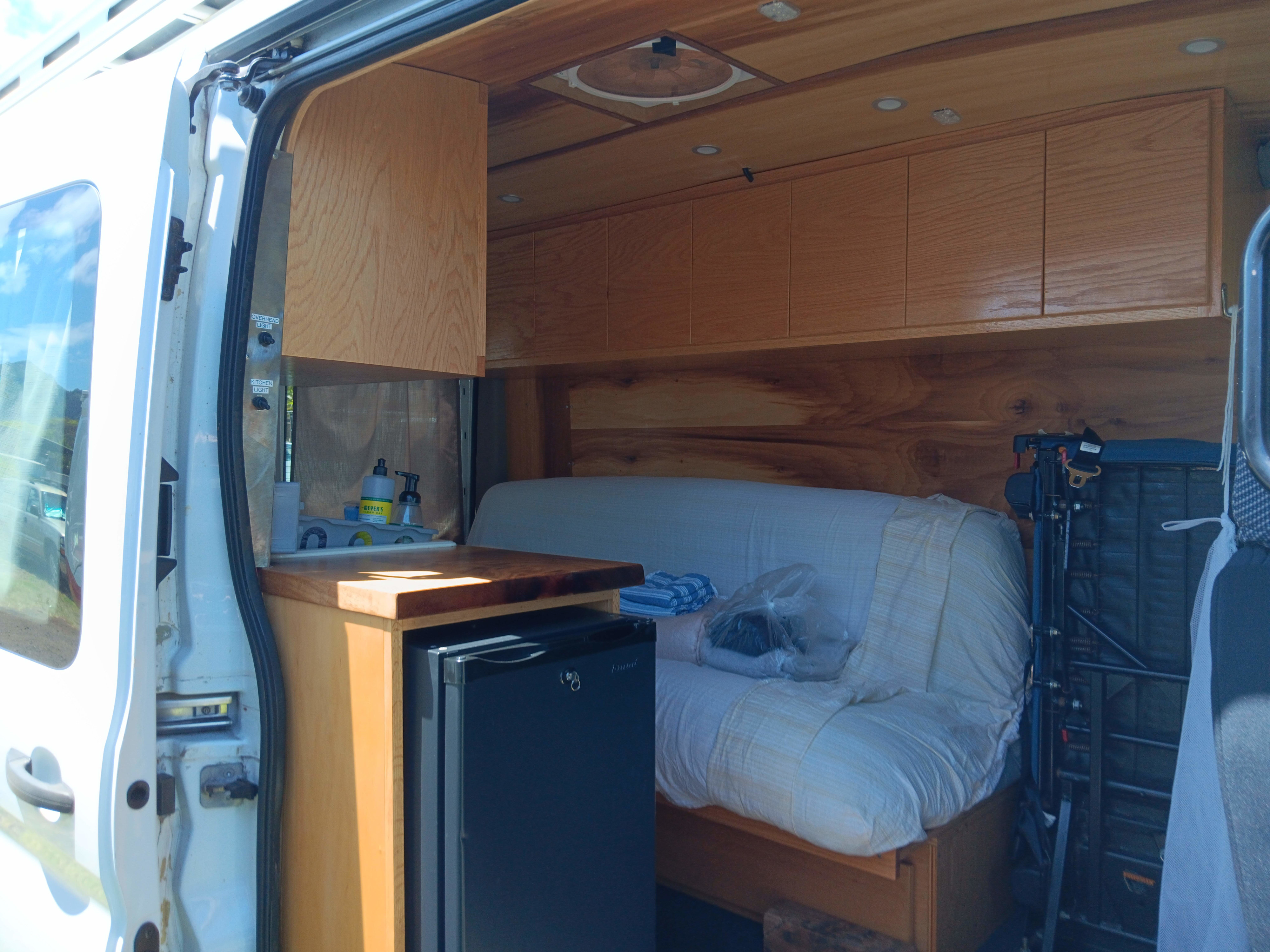 Cozy RV rental interior with custom wood cabinets, countertop kitchen, mini-fridge, and comfortable sofa bed.. Ford Transit 2018