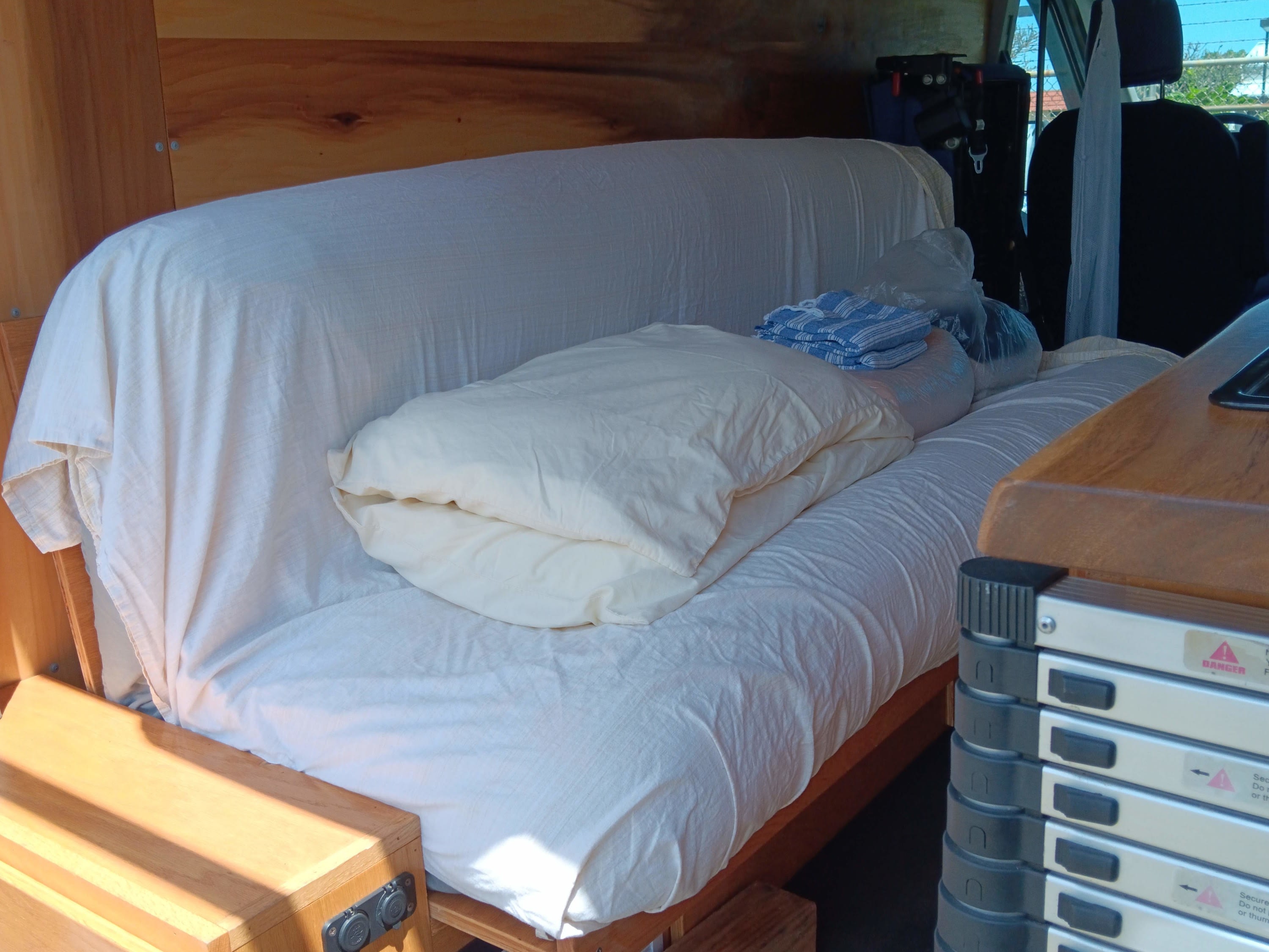 RV rental interior: custom wooden build with a comfortable bench/bed setup, white bedding, and travel essentials.. Ford Transit 2018