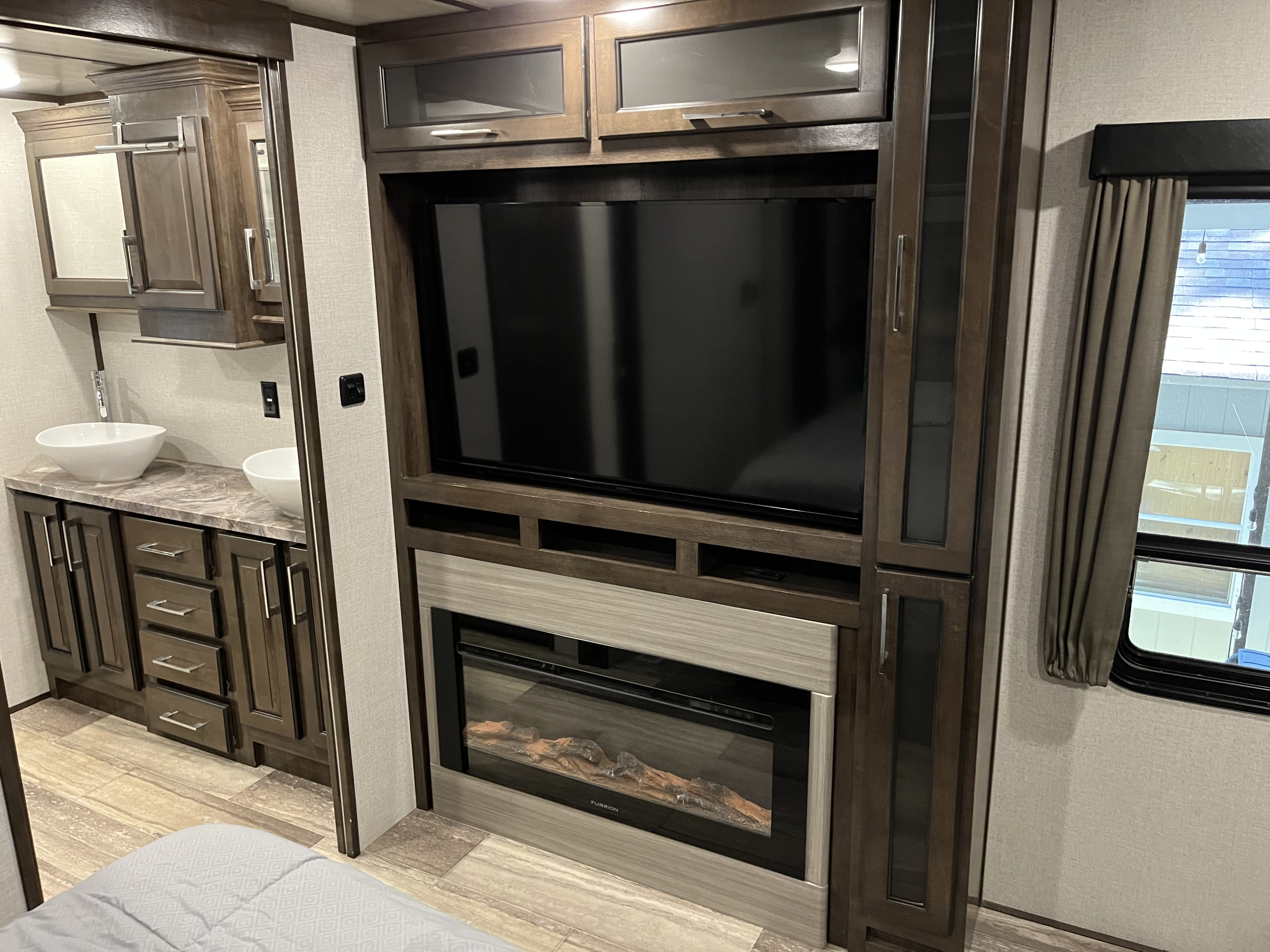 Modern rv rental interior: bedroom with dual vanity, entertainment wall featuring TV & fireplace, and ample dark wood storage.. Dutchmen Yukon 2021