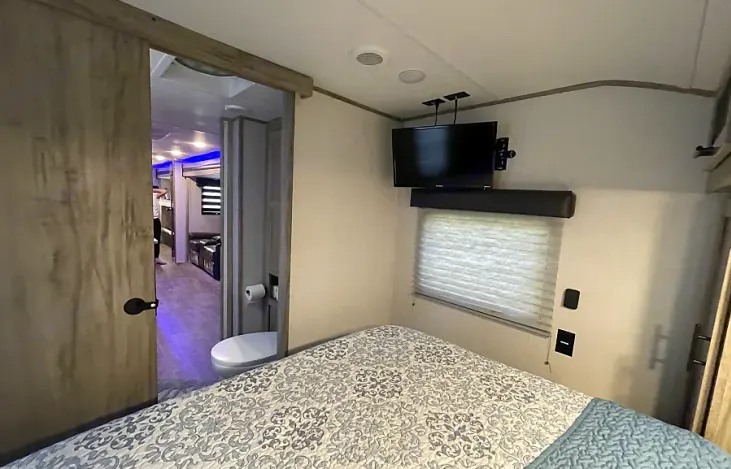 Spacious RV rental interior. Bedroom with TV, window & patterned quilt. Sliding door to compact bathroom & main living space.. Forest River Cherokee Alpha Wolf 2022