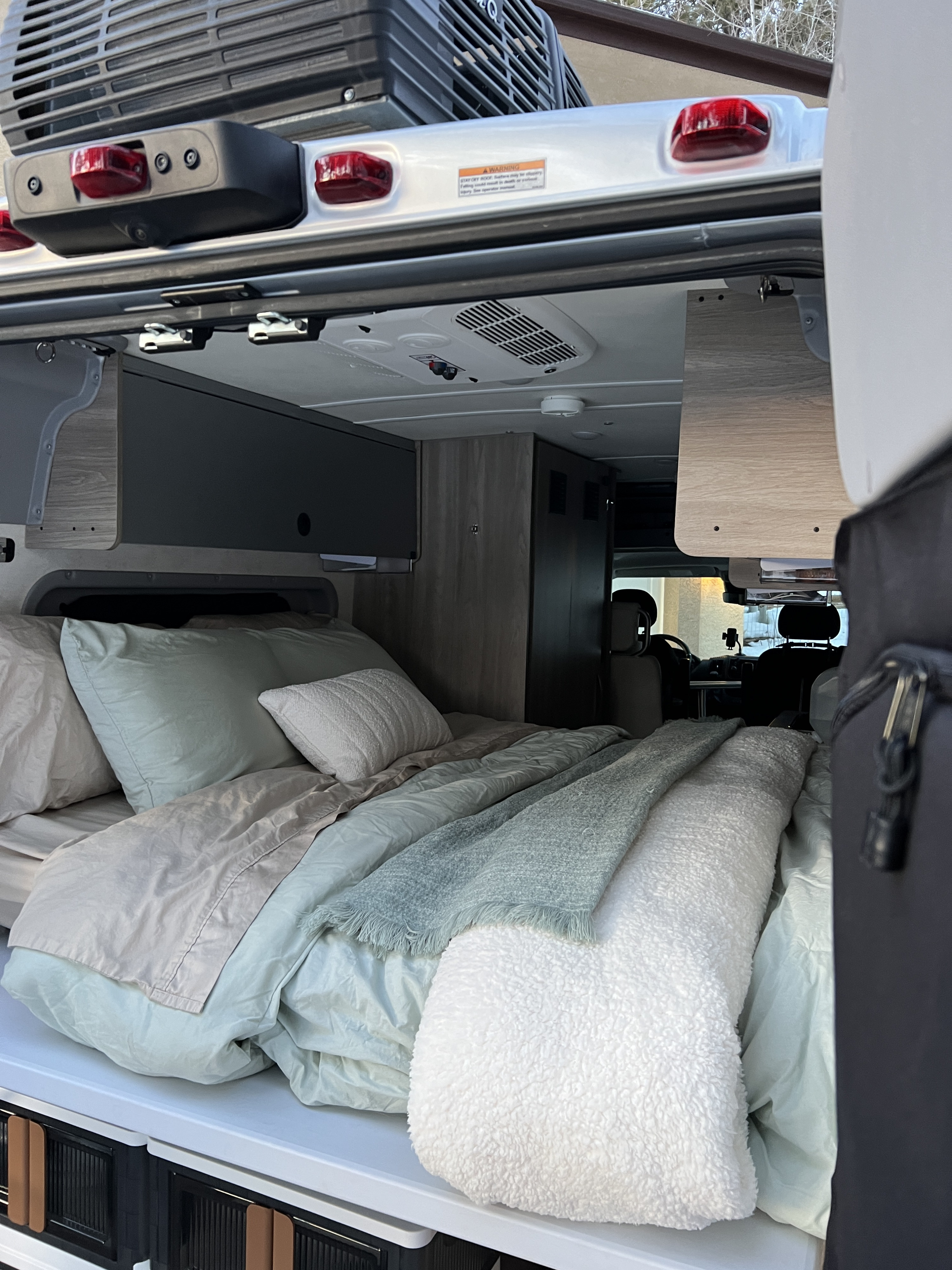 Inside a comfy RV rental: a neat bed with soft bedding, overhead storage, and AC, viewed from the open rear door.. Winnebago Solis 2022