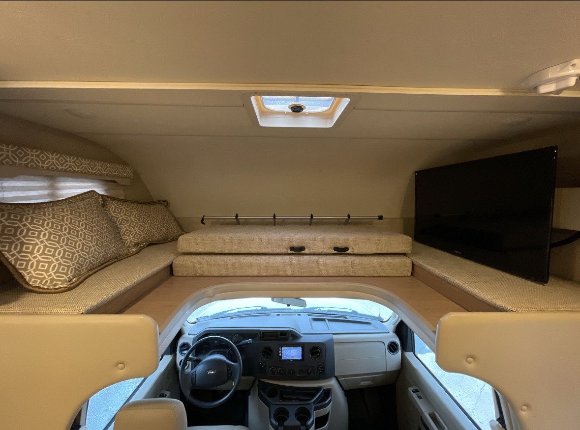 Inside this RV rental: a comfy cab-over bunk bed with patterned pillows, TV, and the driver's cockpit area.. Thor Motor Coach Freedom Elite 2018
