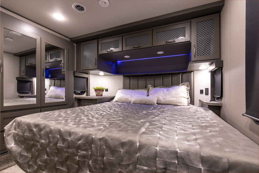 Modern RV rental bedroom with a large bed, gray patterned comforter, overhead cabinets, and blue accent lighting.. Grand Design Momentum M-Class Toy Hauler 2023
