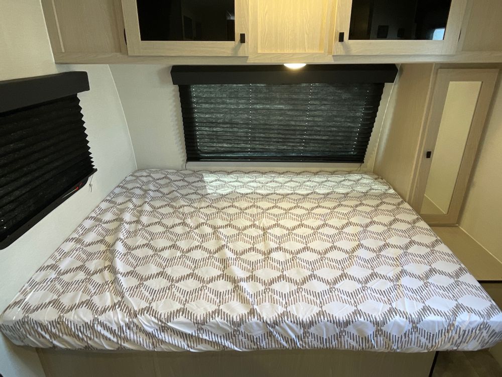 A cozy RV rental bedroom features a large bed with a patterned sheet, overhead cabinets, and window blinds.. Coachmen Apex Nano AAPT186BH 2024