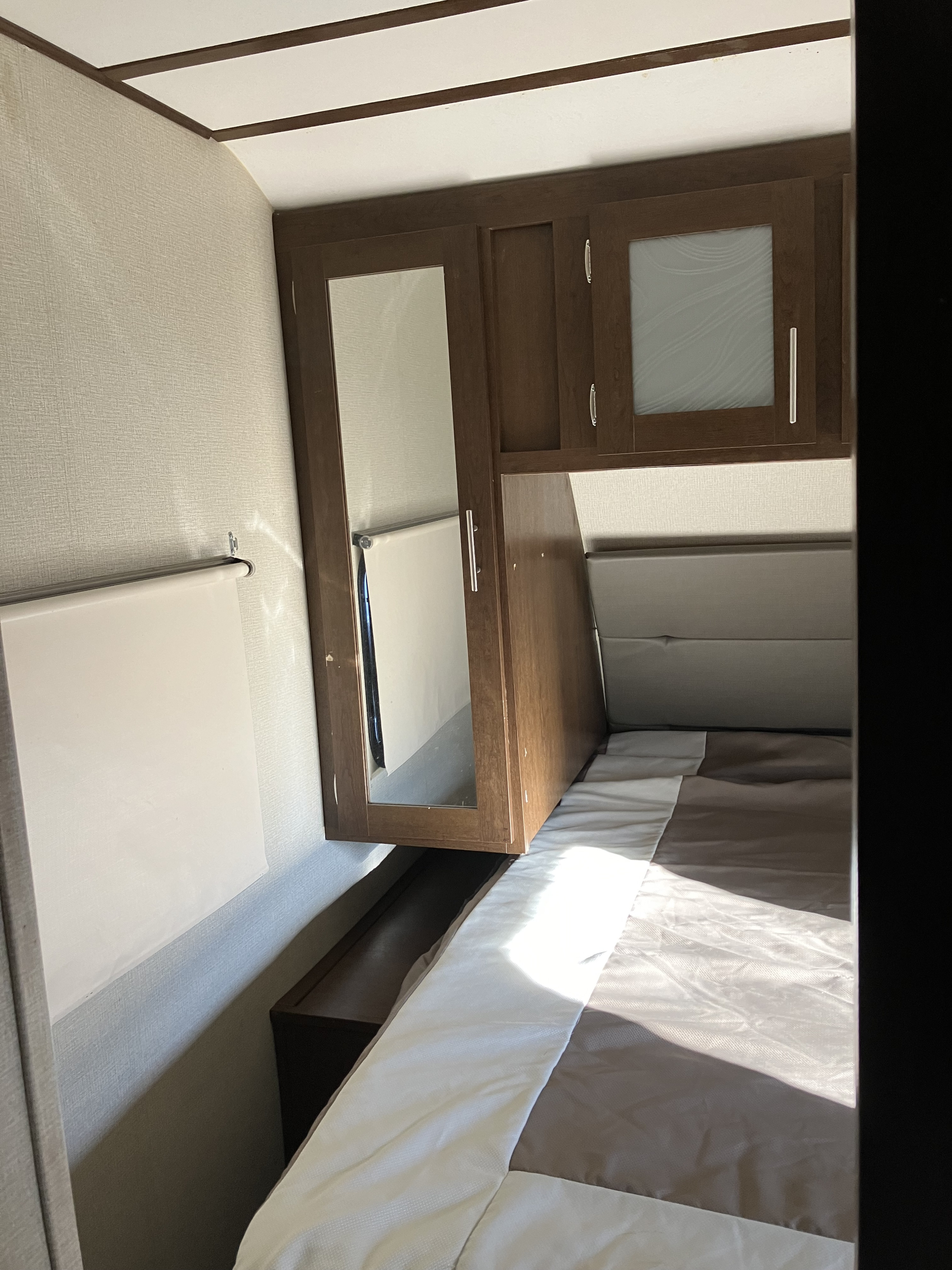 Cozy RV rental bedroom featuring a two-tone bed, overhead wooden storage cabinets with mirror and frosted glass, and a pull-down blind.. Forest River Wildcat 2019