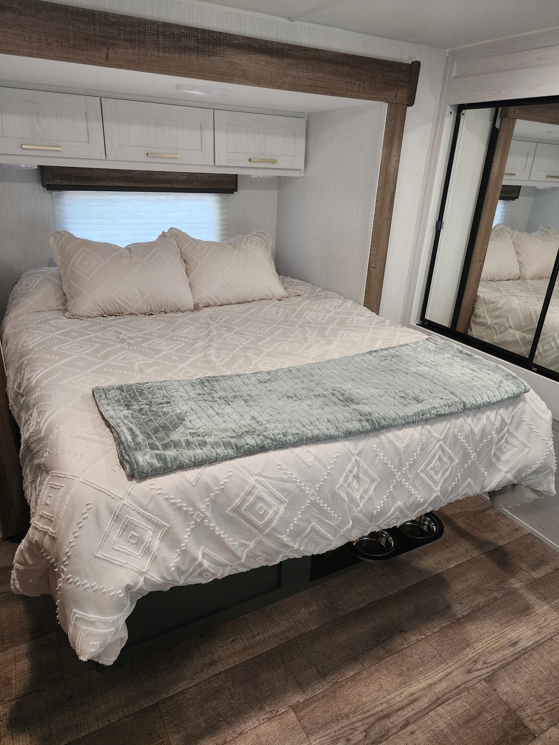 Cozy RV rental bedroom: light-colored bedding with diamond pattern, teal throw, overhead cabinets, and wood-look details.. Heartland RVs Eddie Bauer 2024