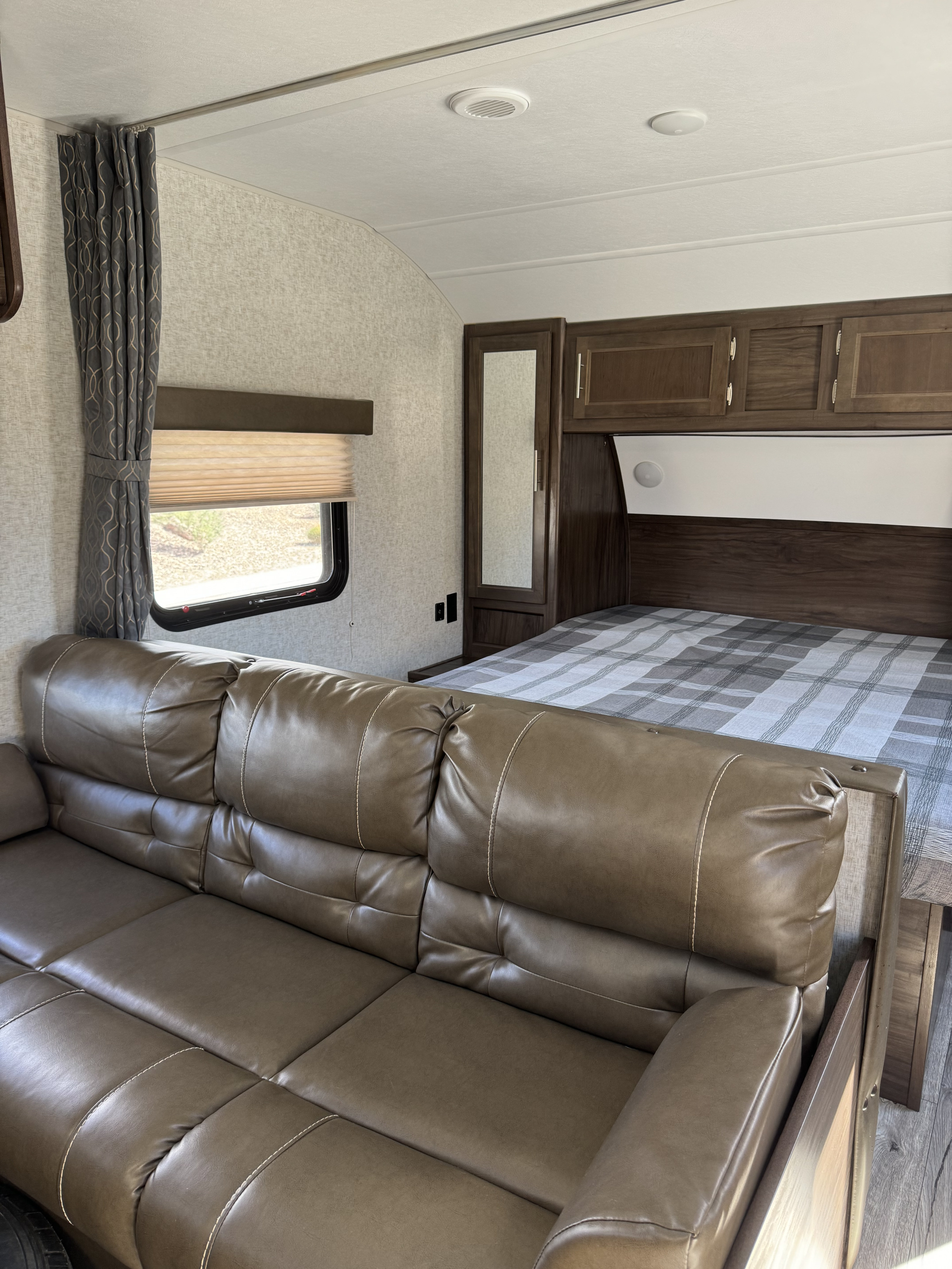 Interior view of an RV rental, showing a cozy brown sofa, window, and compact bedroom with a plaid bed.. Forest River Cherokee 2018