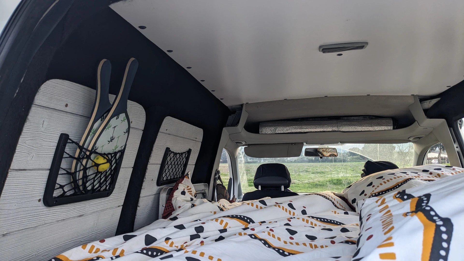 RV rental interior: cozy bed, wall storage with paddles, and a grassy field view from this converted van.. Volkswagen Caddy 2012