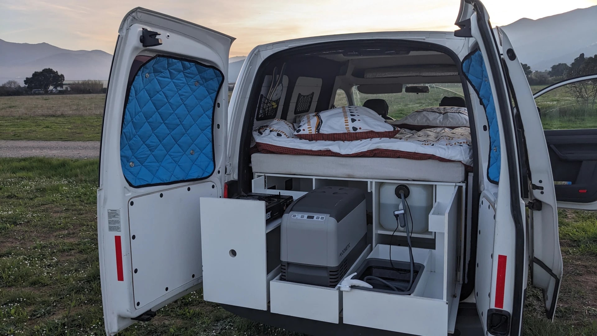 RV rental camper van interior: open doors show a bed, pull-out kitchen with fridge and stove for outdoor living.. Volkswagen Caddy 2012