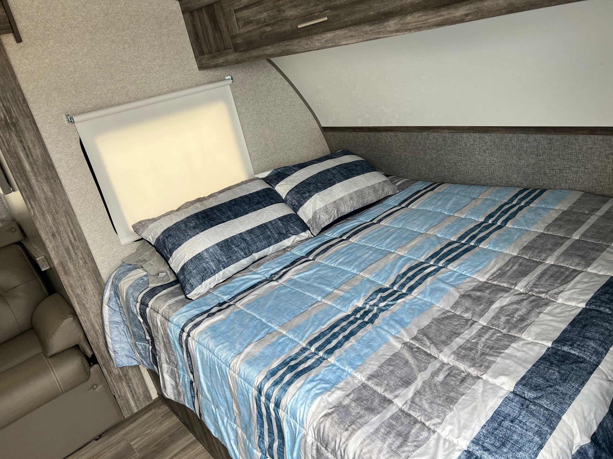 Cozy RV rental bedroom featuring a bed with a blue/grey striped comforter, window shade, and light wood flooring.. Forest River Wildwood FSX Platinum 2020