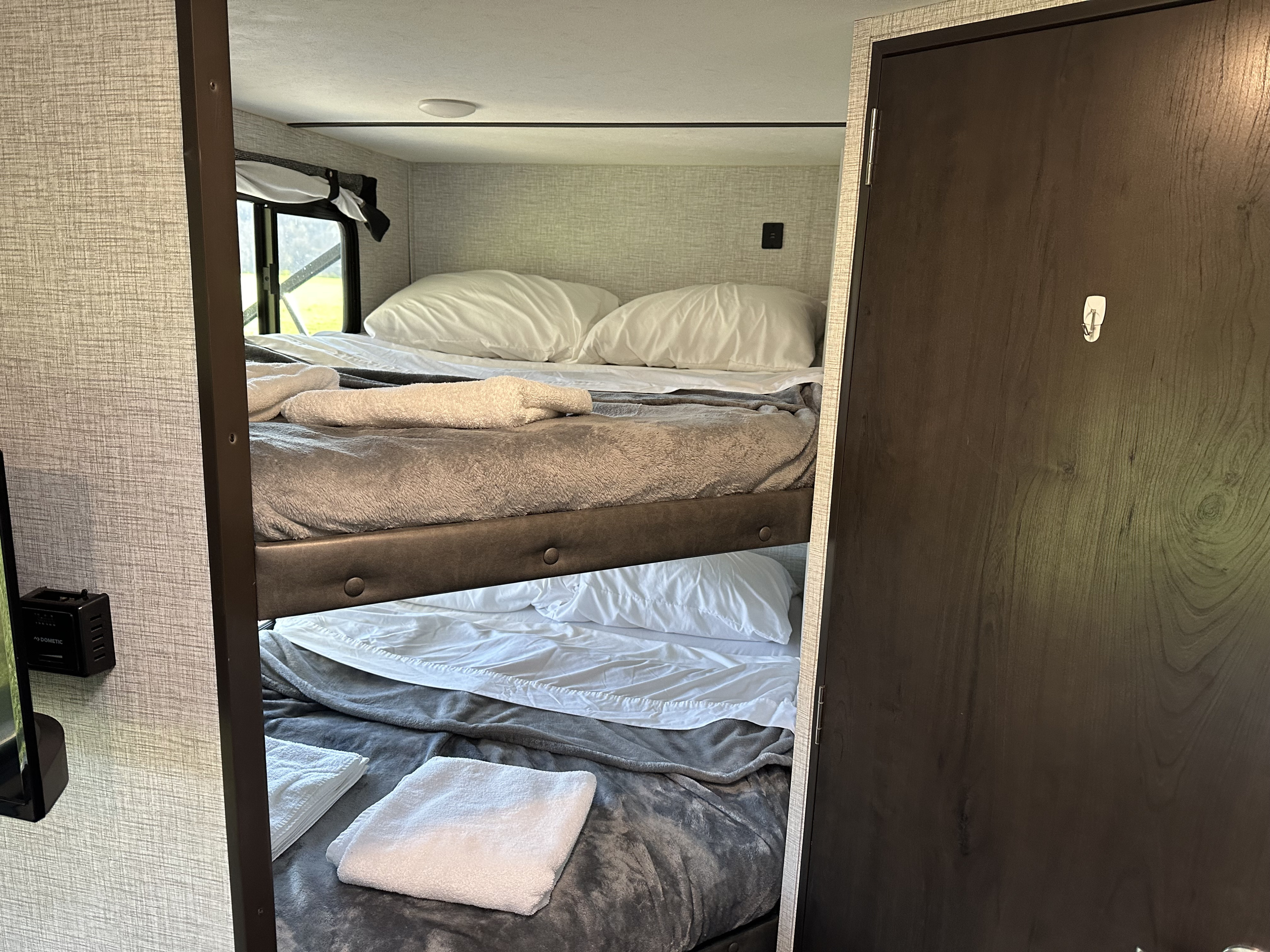 Inside an RV rental: Two tidy bunk beds with white pillows & soft grey blankets. A window shows the green outdoors.. Keystone RV Bullet Crossfire 2021