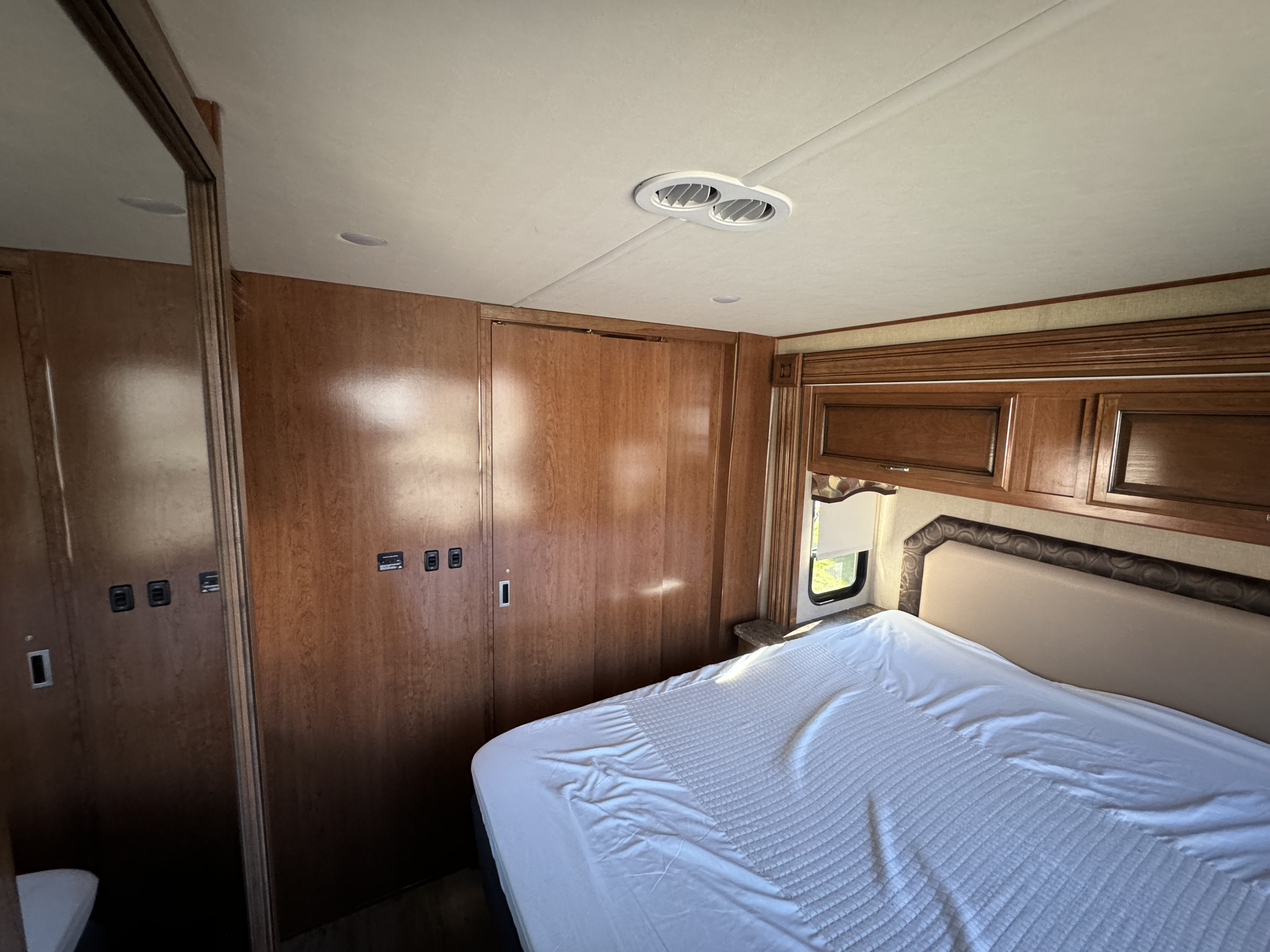 This RV rental's bedroom shows a bed with white linen, wood cabinets, and a window, providing a functional and compact sleeping area.. Flair-Fleetwood Flair-Fleetwood Motorhome 2017