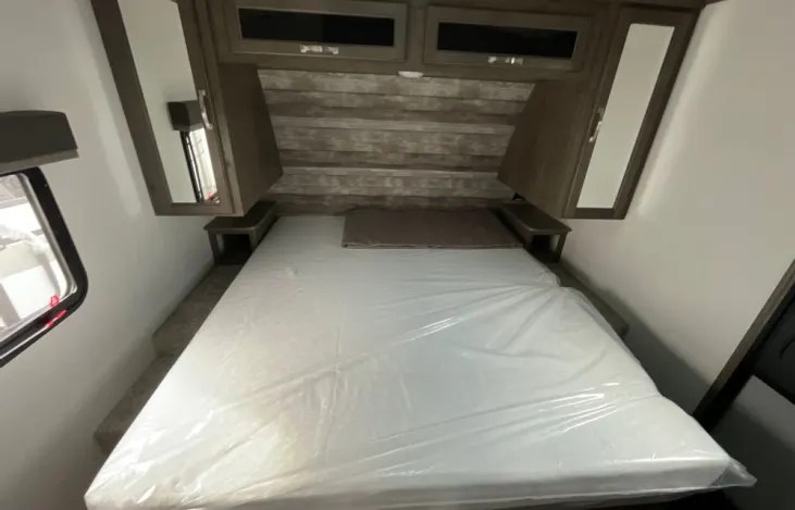 Inside an RV rental: a compact bedroom features a bed with a new mattress, wooden headboard, overhead cabinets, and side mirrors.. Keystone RV Cougar 2023