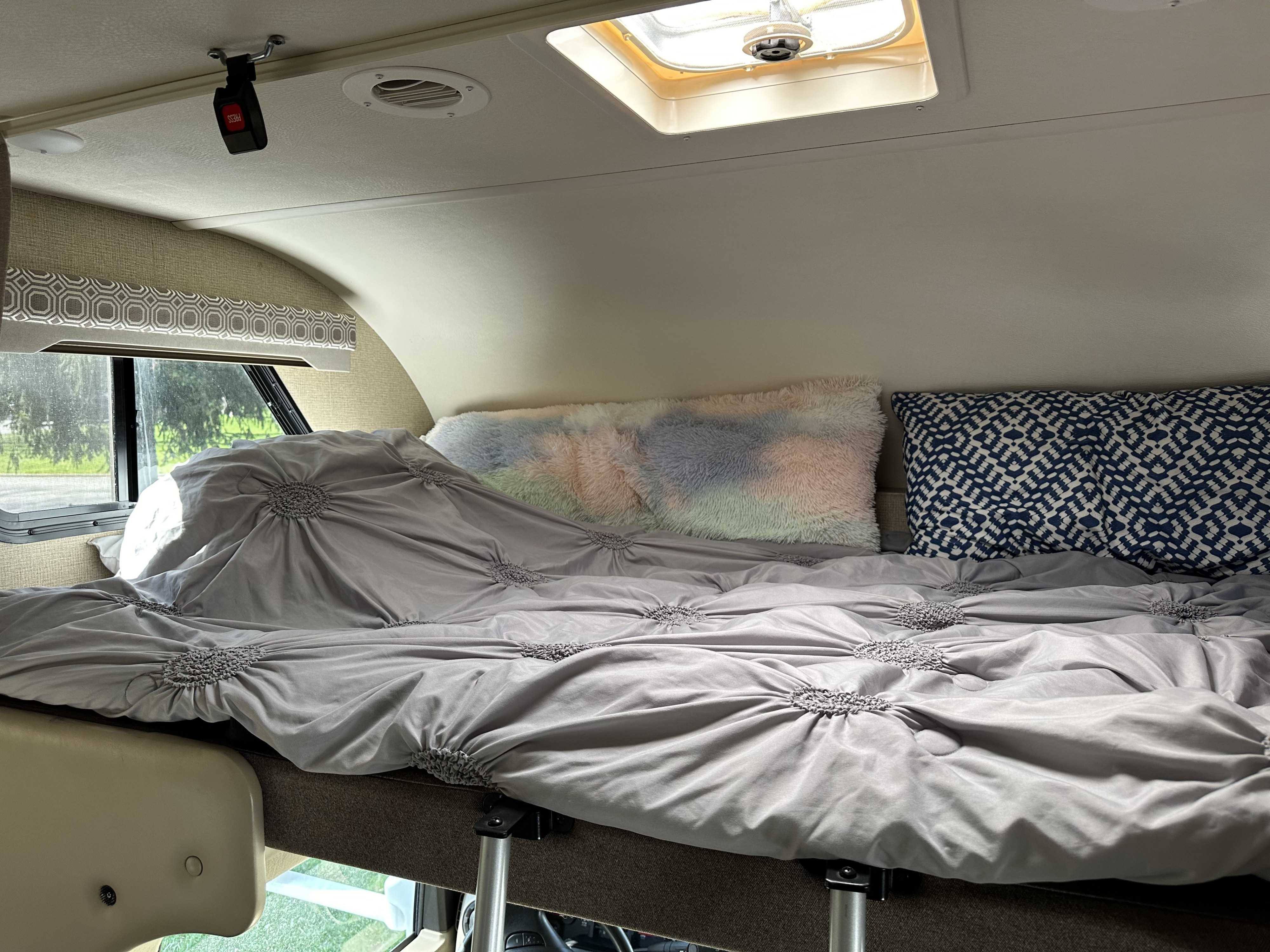 Cozy cab-over bunk bed in an RV rental, featuring grey bedding, colorful pillows, a window, and ceiling fixtures.. Thor Four Winds 2019