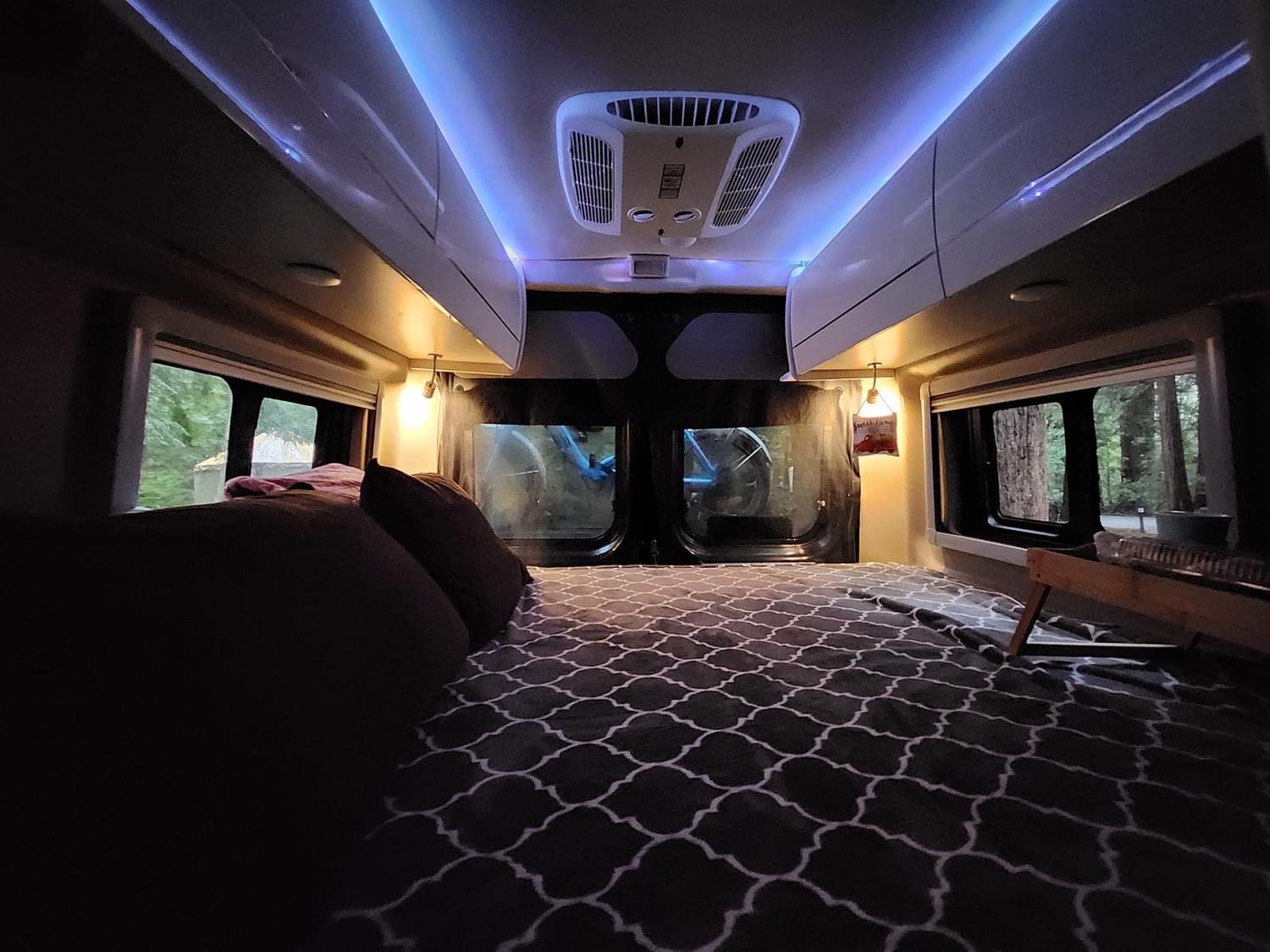 Inside a comfy RV rental: bed with patterned comforter, blue ambient lighting, and large windows revealing a peaceful forest.. Thor Tellaro 2023