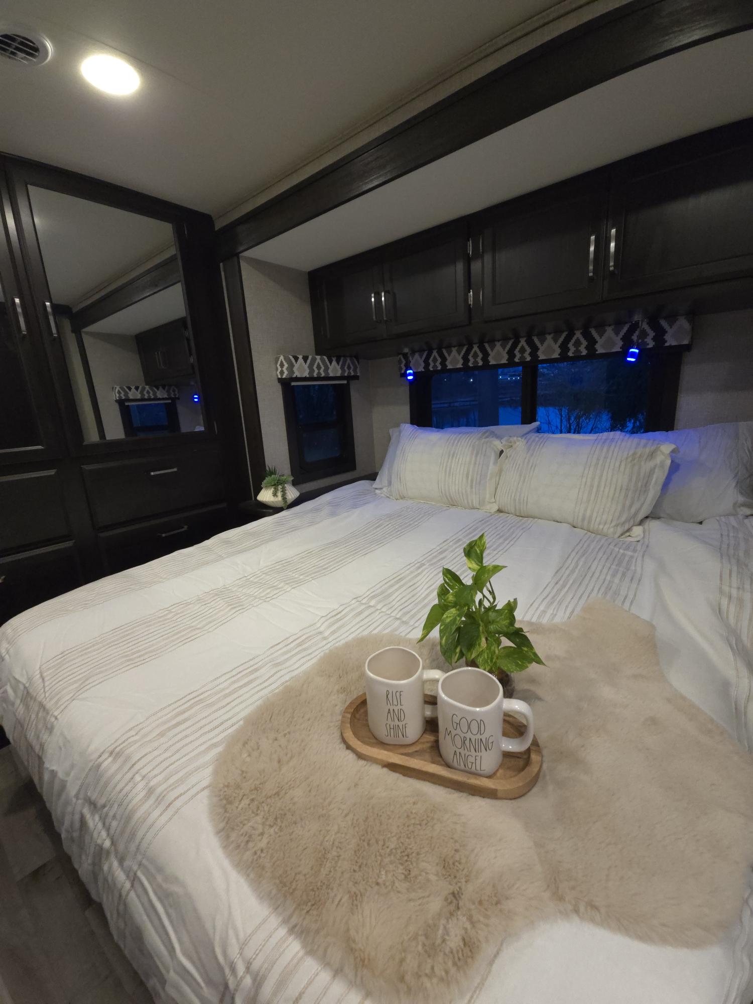 Cozy RV rental bedroom with comfy striped bedding, coffee mugs on a fur throw, and dark cabinets. Ideal for travel comfort.. Jayco Alante 2023