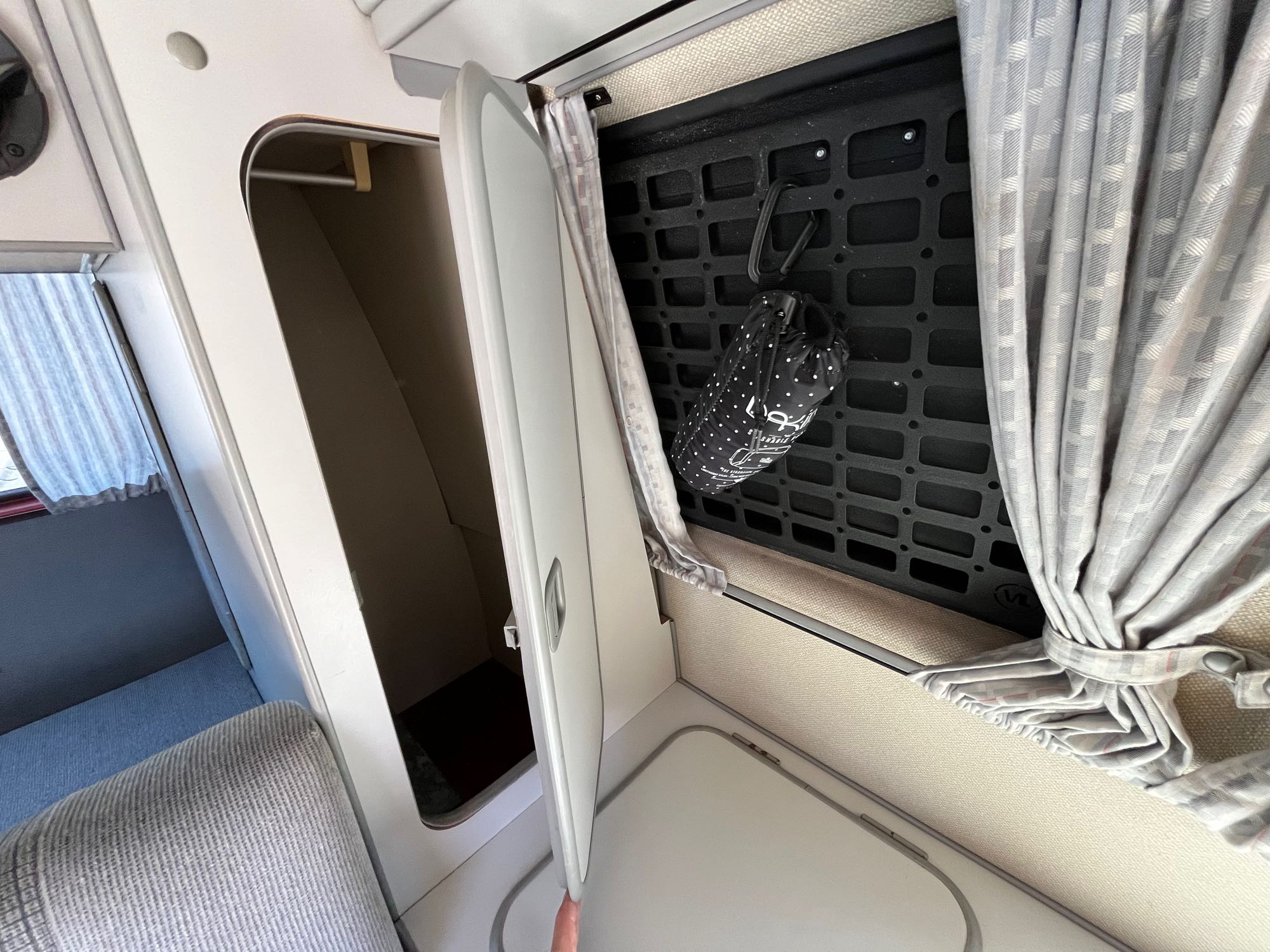 RV rental interior with open closet, cabinet door, and organized side panel displaying a hanging bag and tied curtains.. Volkswagen Westfalia 1987