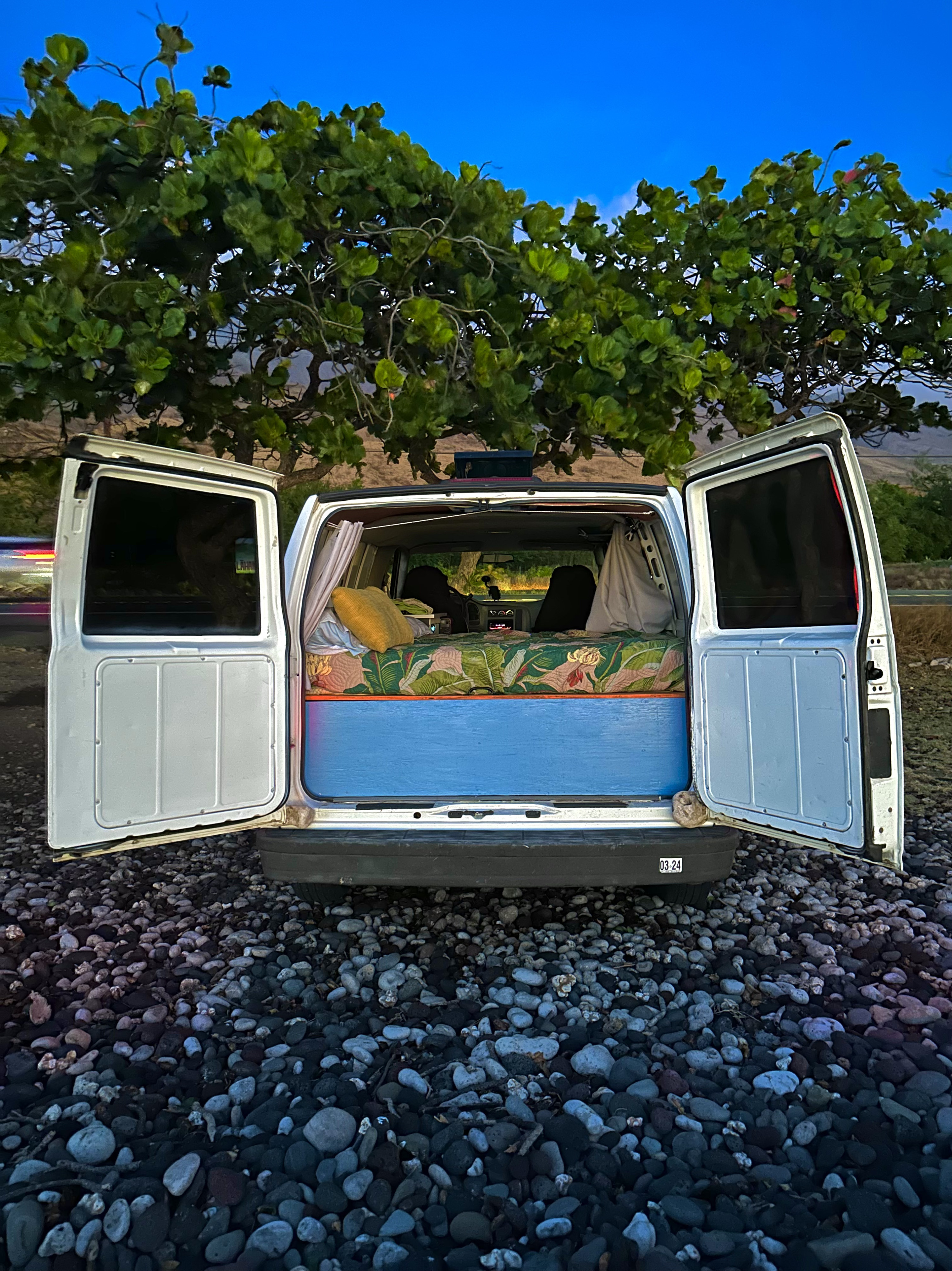An RV rental camper van with open rear doors reveals a cozy bed, parked on a rocky beach at dusk with lush green trees.. Chevrolet Other 2005