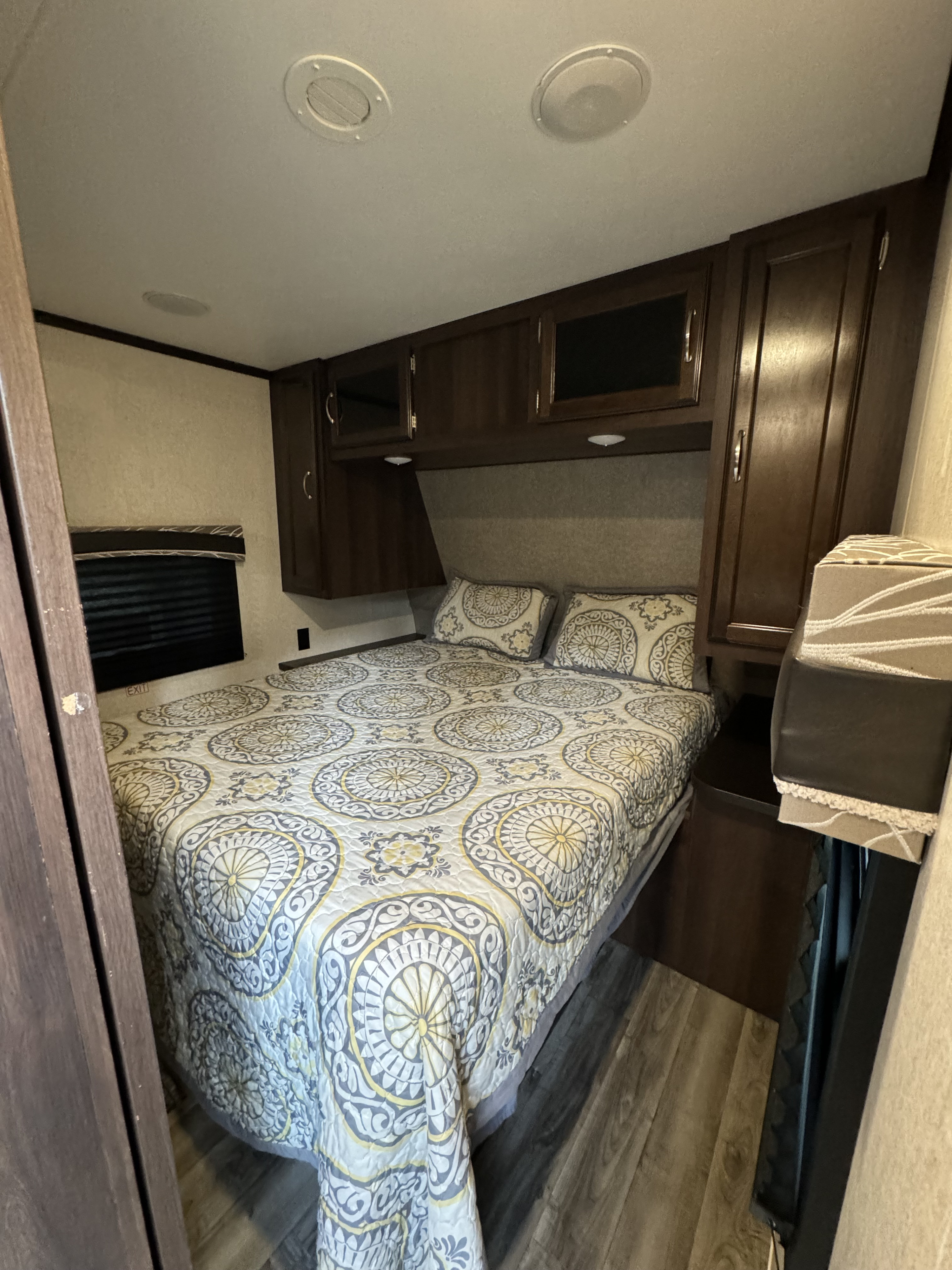 Cozy RV rental bedroom featuring a queen bed with a patterned comforter, dark wood overhead storage, and a window.. Jayco Jay Flight 2018