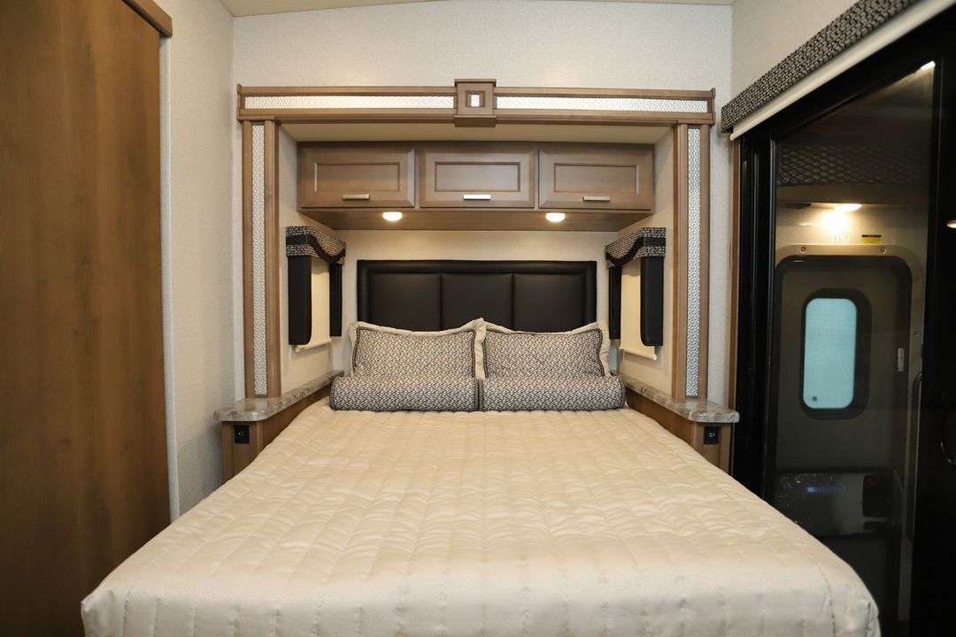 View of a comfortable RV rental bedroom, featuring a queen bed, overhead cabinets, padded headboard, and an exit door.. Thor Outlaw 2023