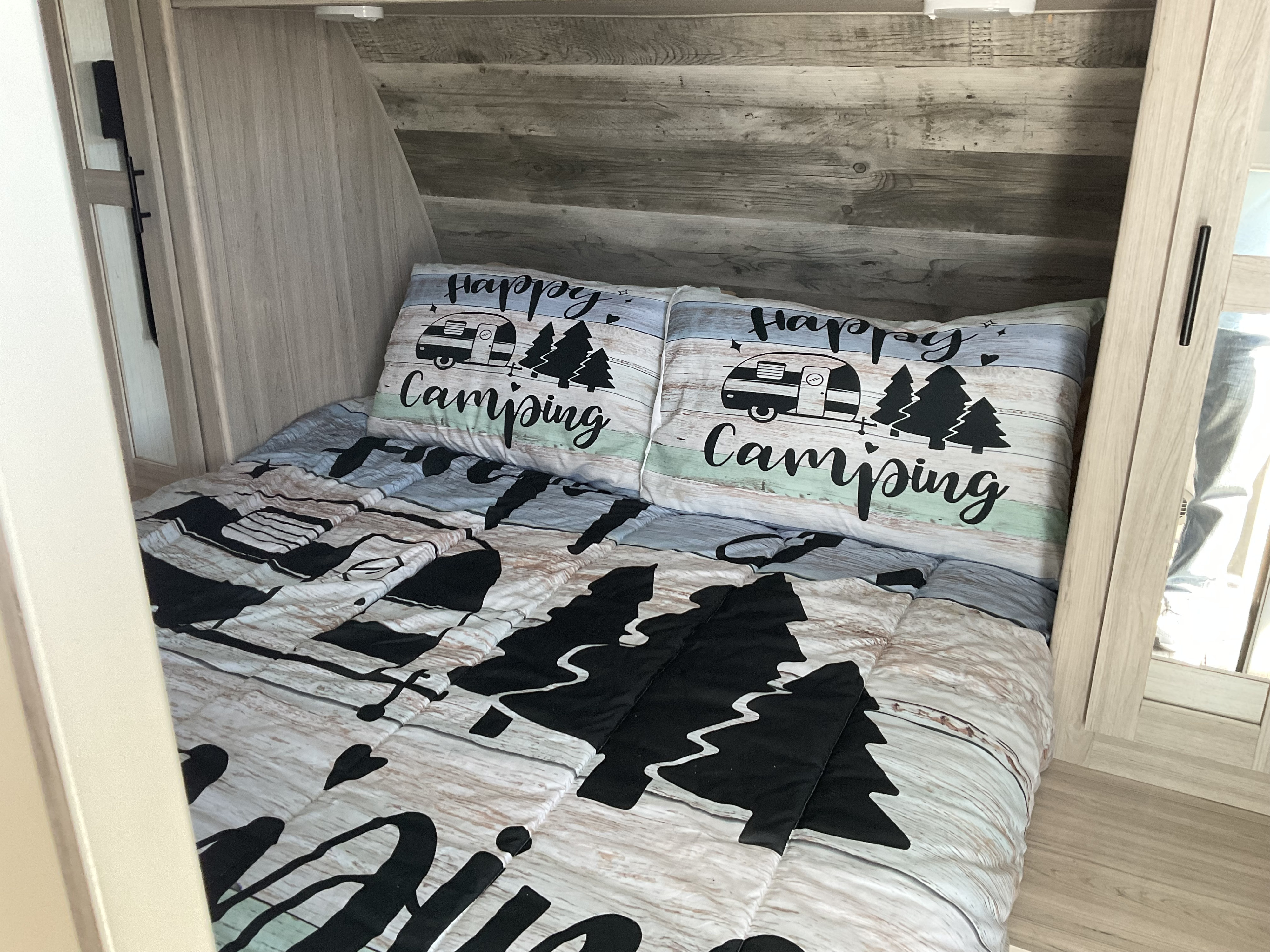 Cozy RV rental bedroom with "Happy Camping" bedding, wood-paneled walls, and mirrored closet door.. Heartland RVs Sundance Ultra Lite 2022